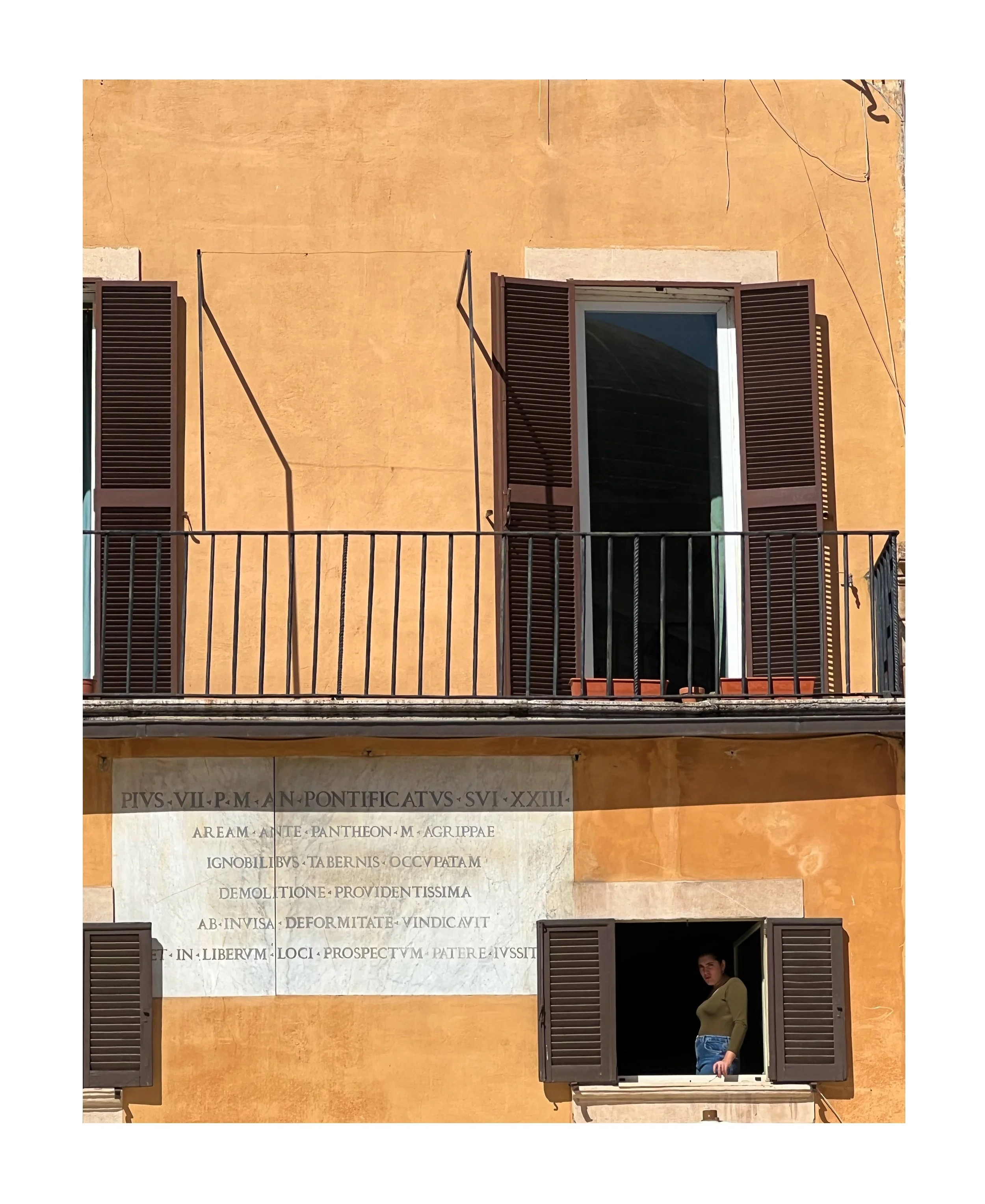 Bâtiment coloré avec deux fenêtres dotées de volets en bois, une avec une personne regardant dehors, et une plaque commémorative en dessous.