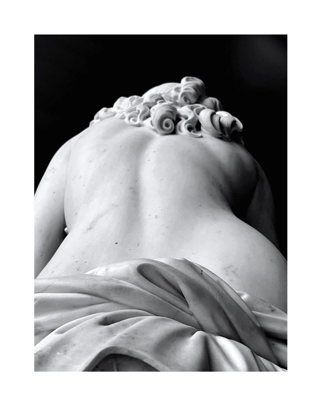 Statue en marbre blanc vue de dessous, avec des détails de cheveux bouclés et de draperie.