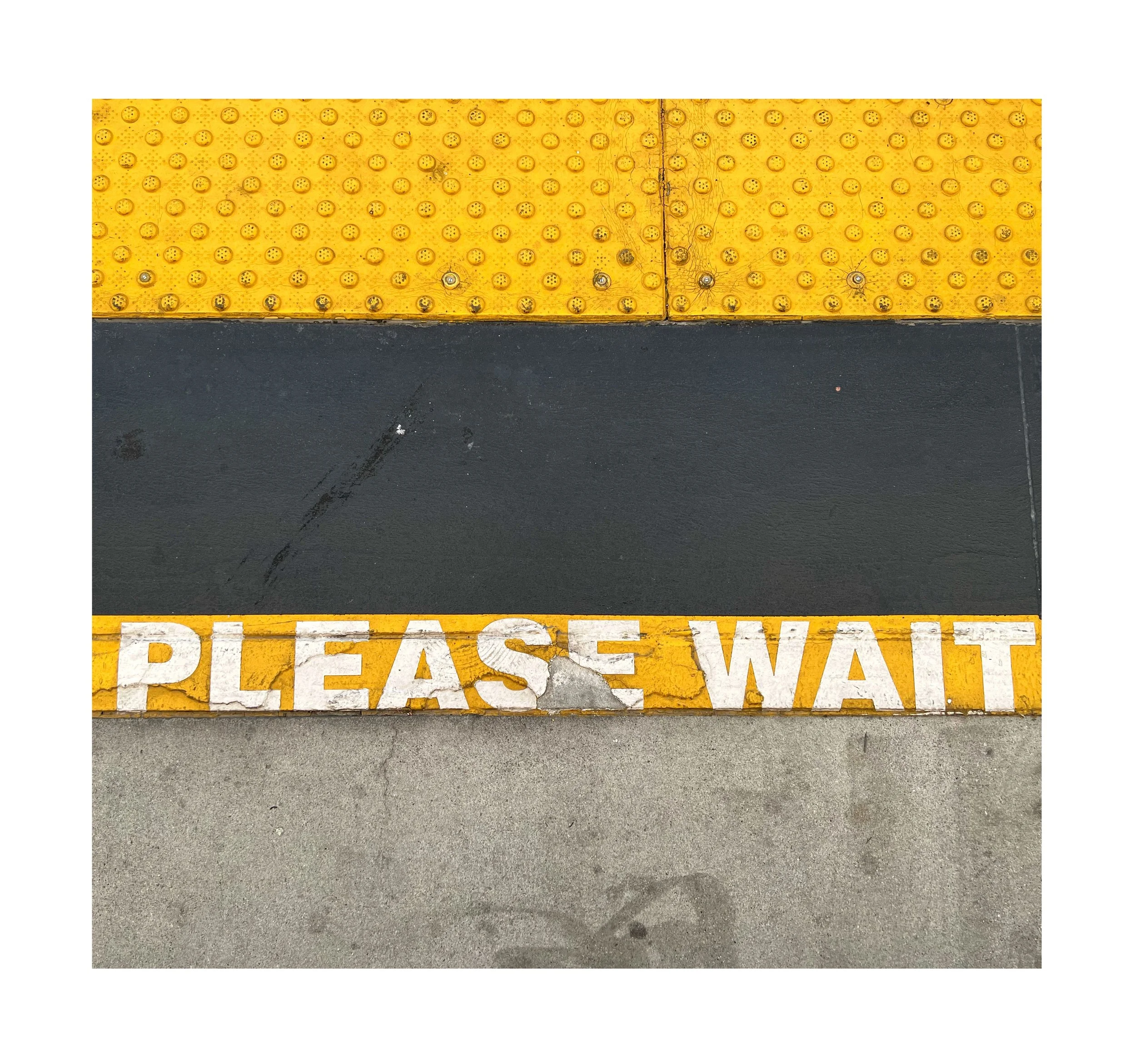 Marquage au sol avec le texte 'PLEASE WAIT' en blanc sur fond jaune, séparé par une ligne noire et une zone grise, avec une surface de sol en béton.