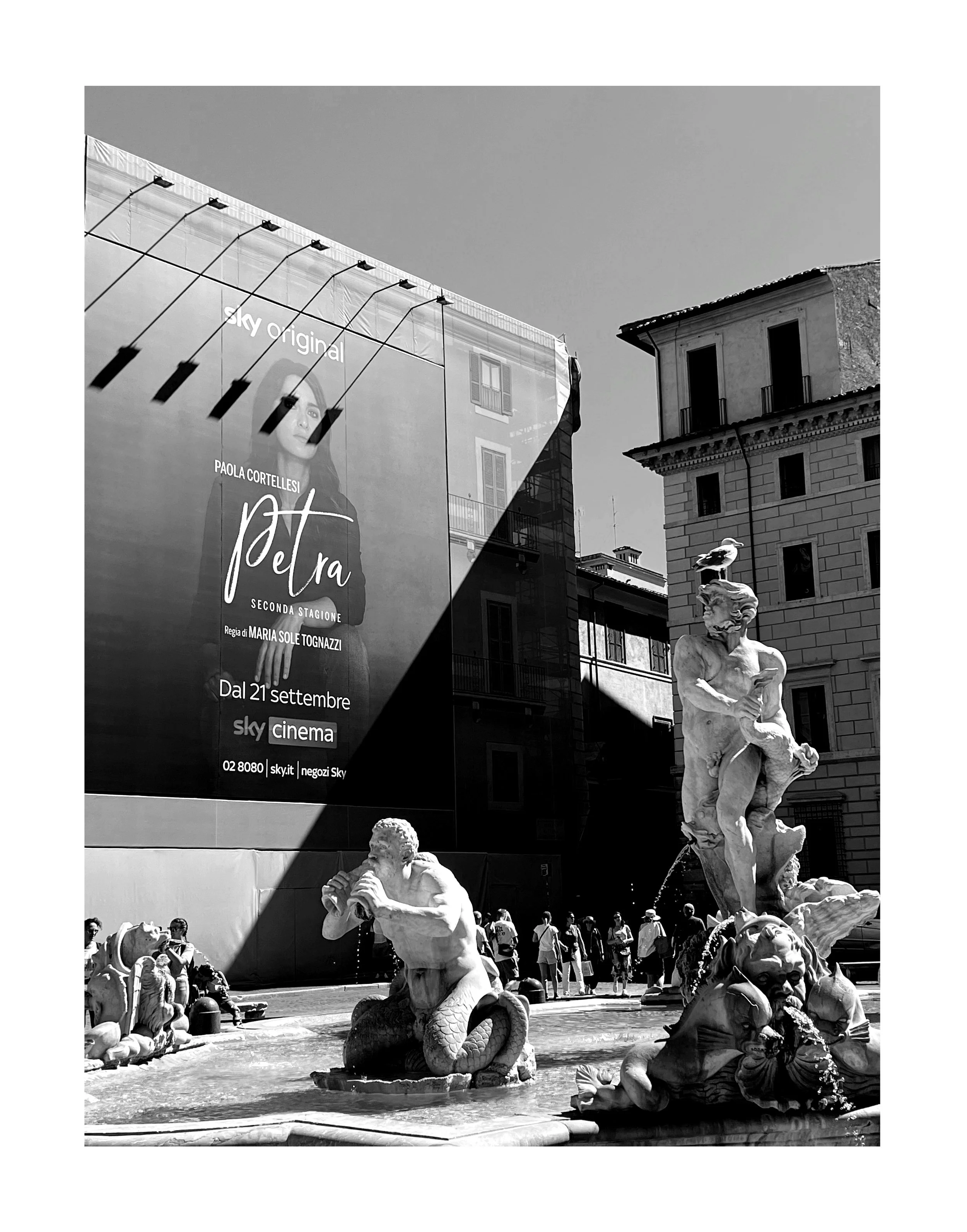 Une place urbaine avec une fontaine de sculptures classiques, entourée de bâtiments anciens et modernes. Un panneau publicitaire pour la série "Petra" est visible à gauche.