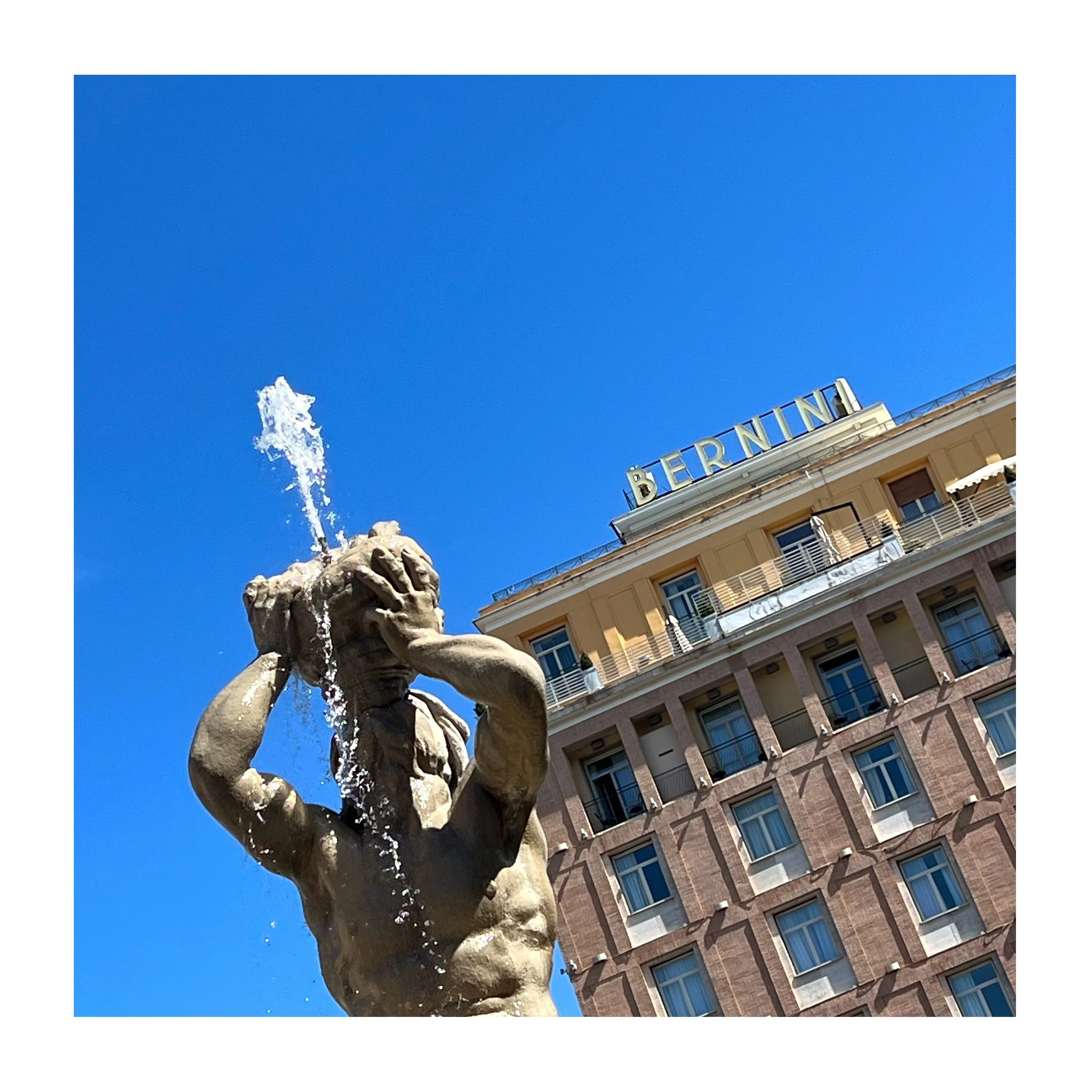Une fontaine en pierre représentant un homme tenant une tête d'animal, avec de l'eau jaillissant de la tête. En arrière-plan, un bâtiment avec un signe lumineux 'BERNINI' sous un ciel bleu clair.