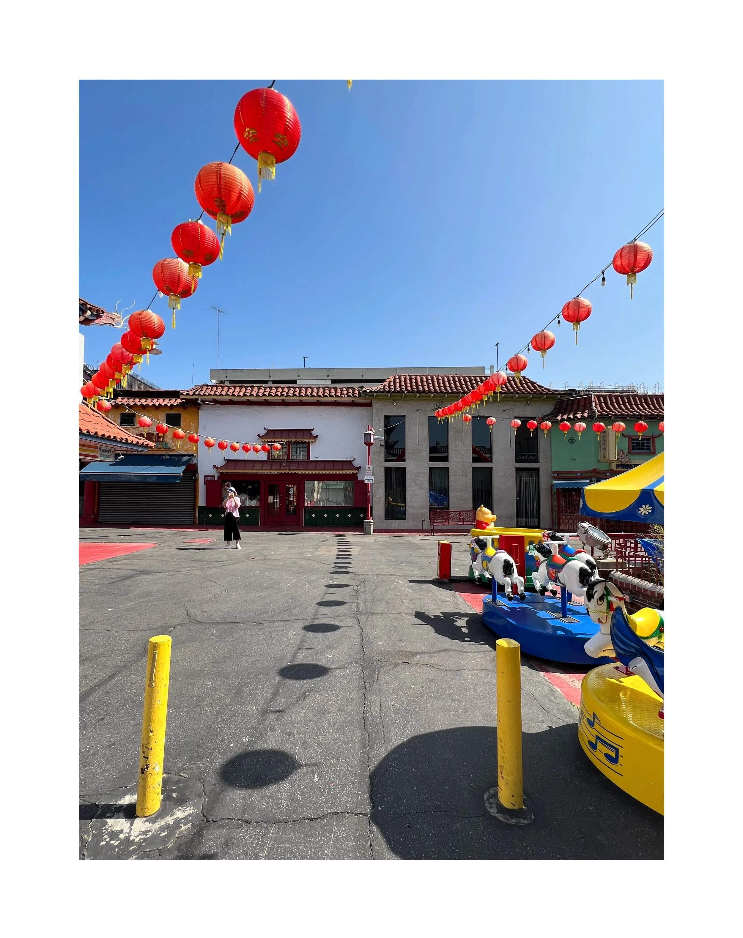 Une place avec des lanternes rouges suspendues au-dessus, des bâtiments colorés et des manèges pour enfants, sous un ciel clair.