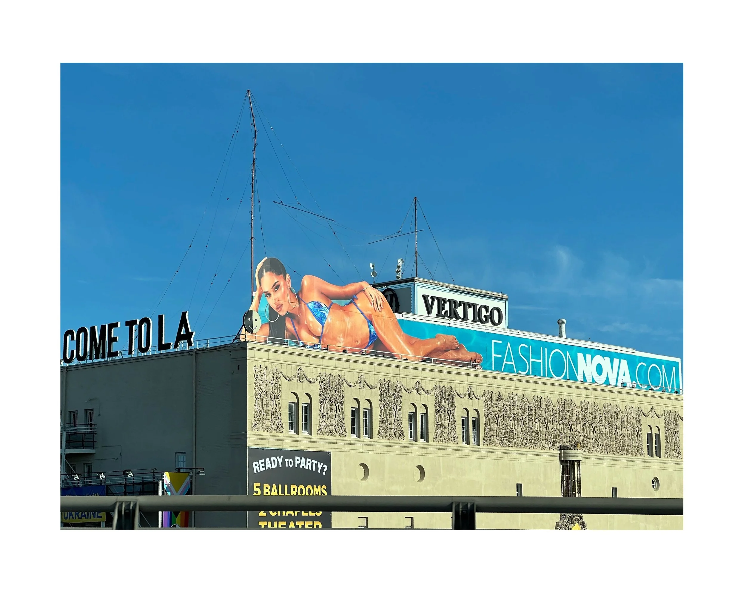 Une grande affiche publicitaire représentant une femme en maillot de bain bleu, allongée sur le toit d'un bâtiment. Le fond du ciel est bleu clair. Il y a des panneaux avec des textes promotionnels, y compris 'COME TO LA', 'VERTIGO', et 'FASHIONNOVA.