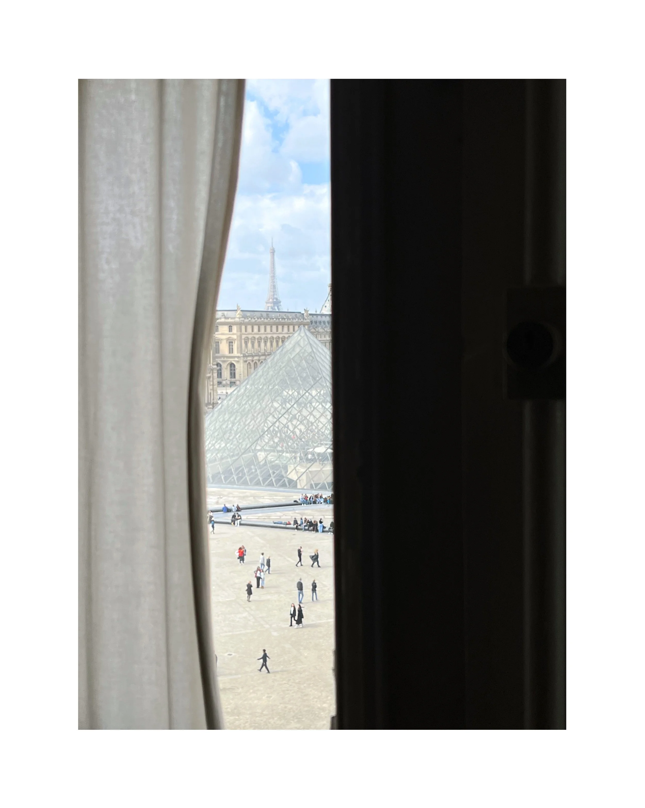 Vue de Paris avec la pyramide du Louvre et la Tour Eiffel en arrière-plan, vue à travers une fenêtre encadrée par un rideau blanc et un volet noir.