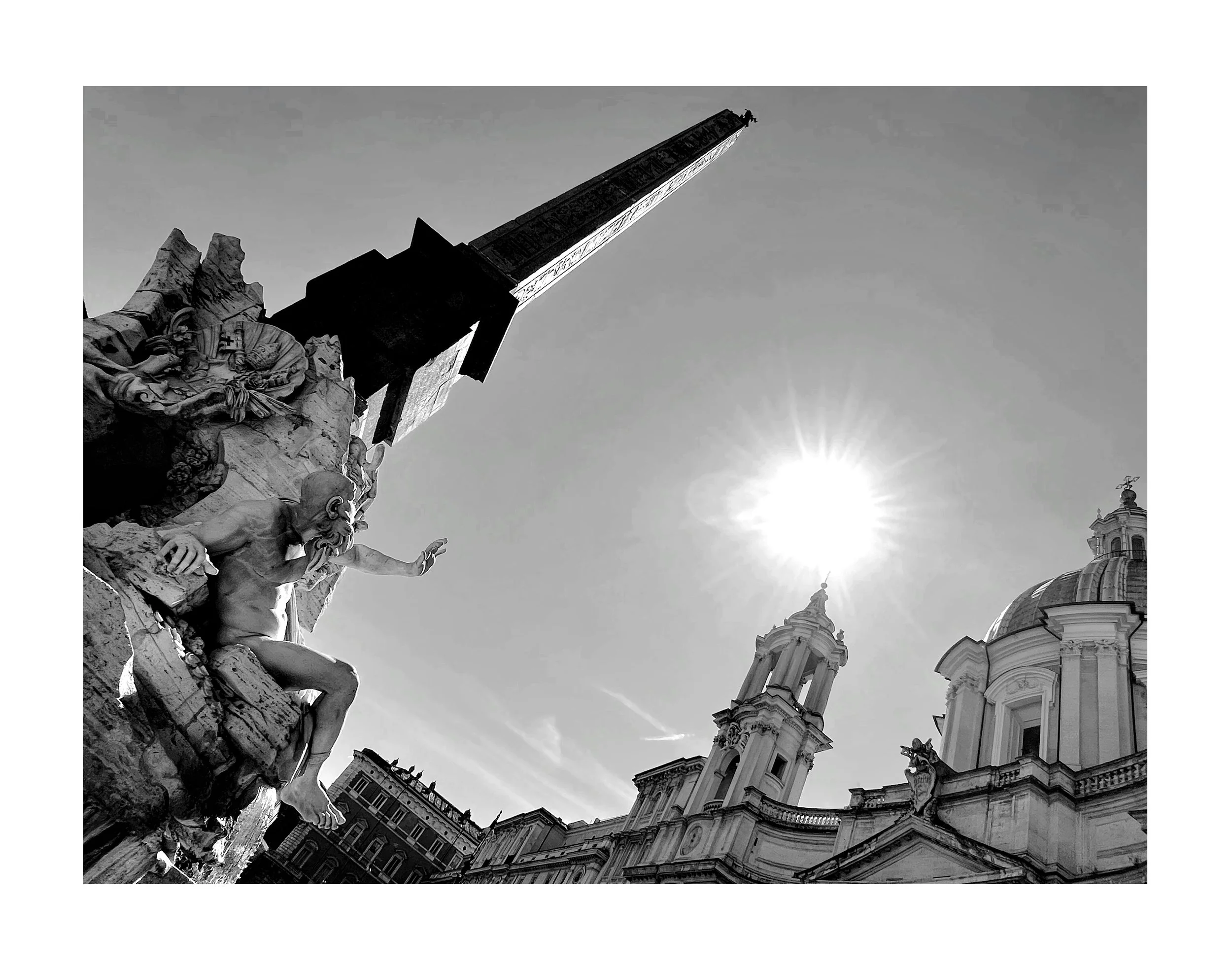 Une photographie en noir et blanc d'une place avec une sculpture baroque, deux clochers et un dôme, avec le soleil brillant haut dans le ciel.