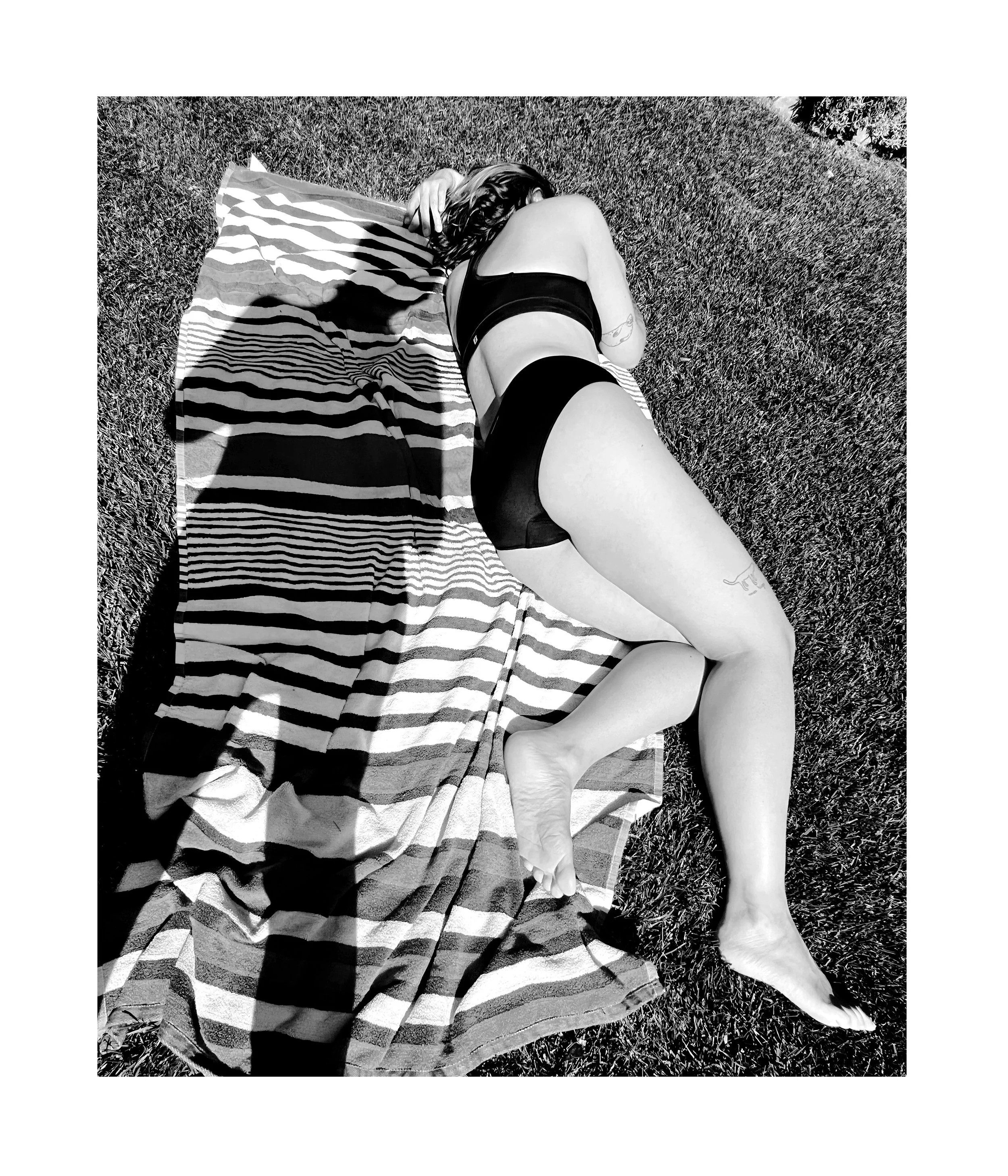 Femme allongée sur une serviette de plage en train de bronzer, vue de dos, en bikini noir, surherbe.