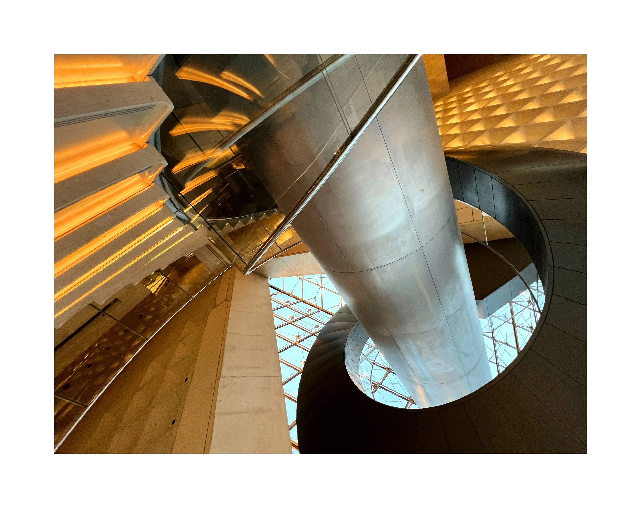 Vue en intérieur d'une architecture moderne avec un escalier tournant et une large conduite de ventilation en métal, entourée de murs en pierre, avec un plafond en verre.