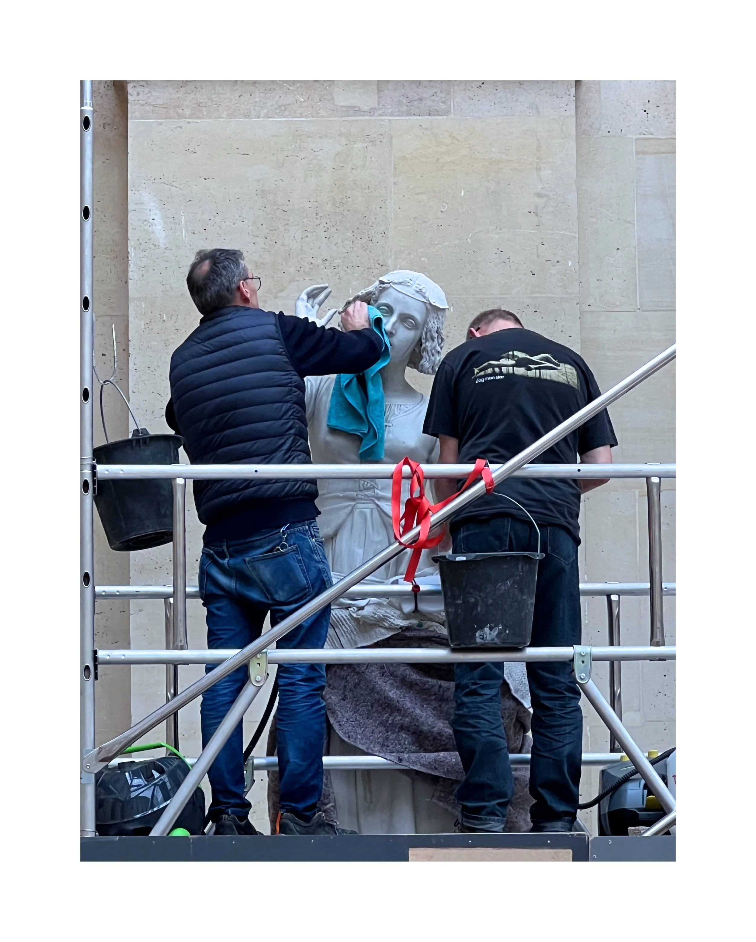 Deux hommes restaurent une sculpture en marbre d'une femme, en utilisant des outils et une échelle, contre un mur en pierre beige.