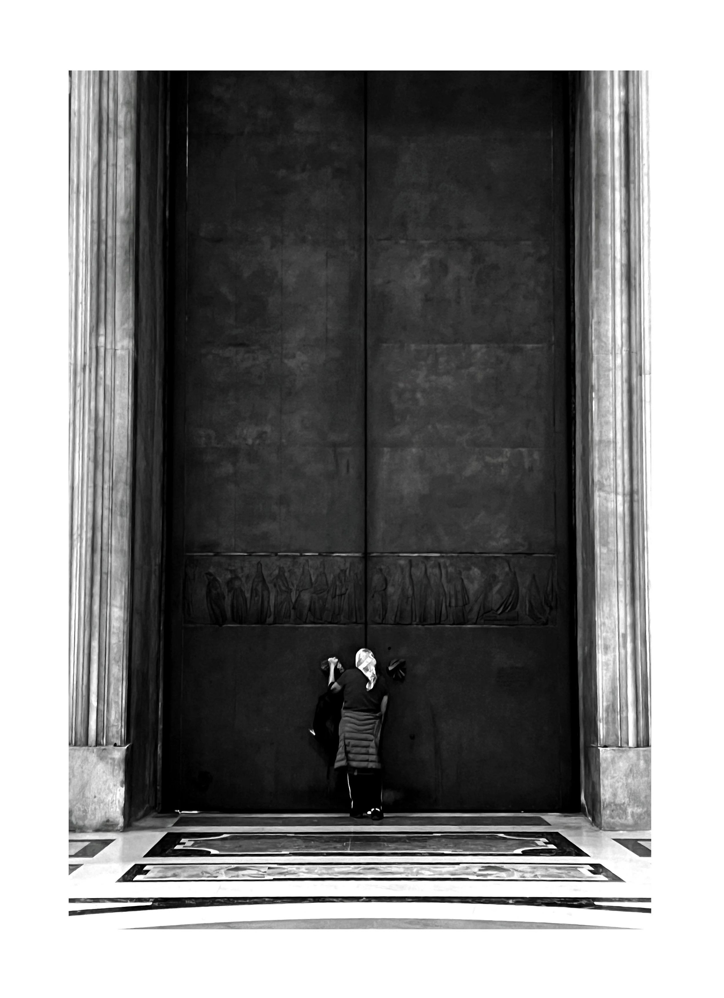 Deux personnes devant une grande porte noire en métal avec des colonnes en pierre de chaque côté, dans un hall architecturale.