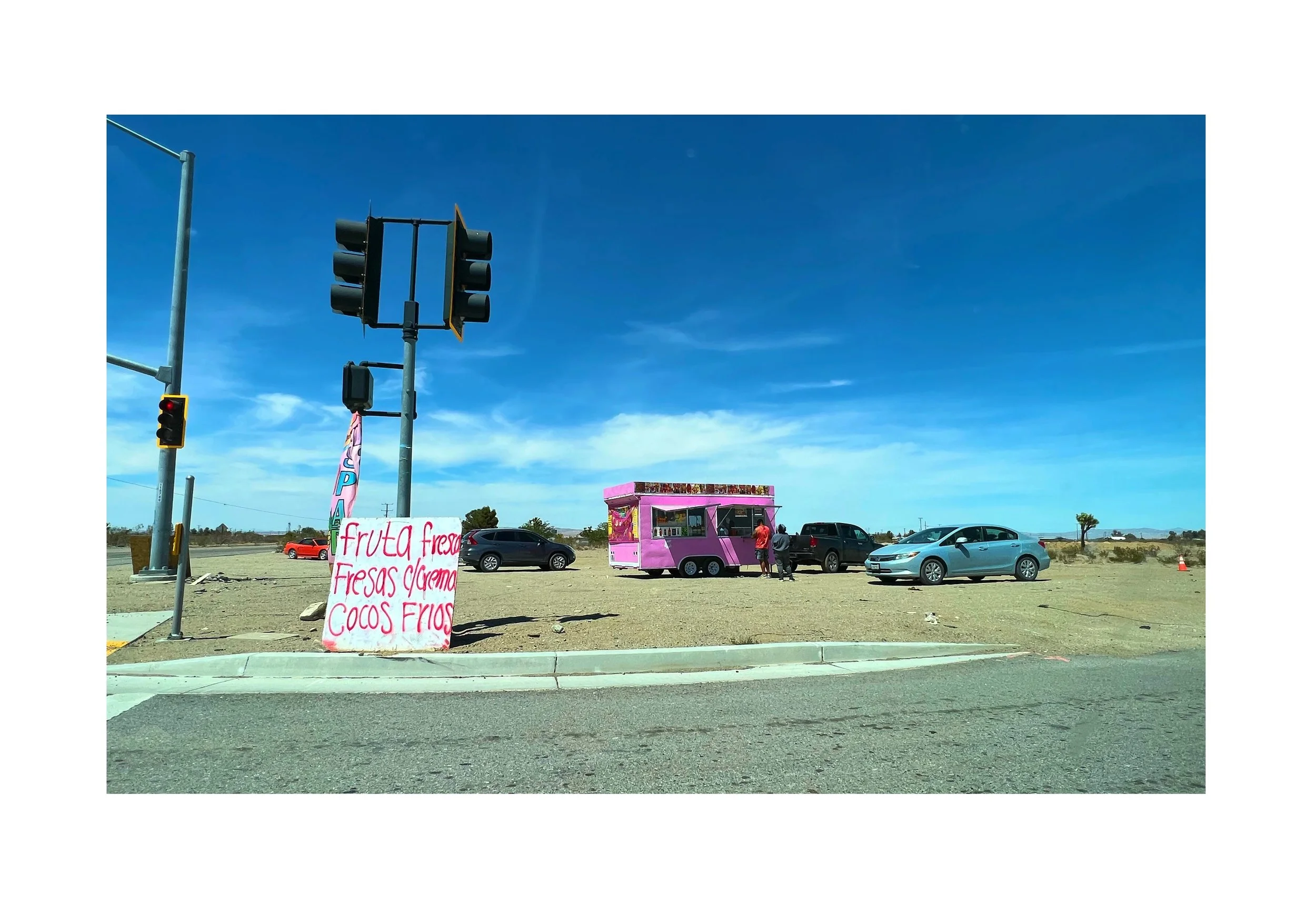 Un stand de vente de fruits en plein air avec un panneau écrit à la main, une caravane rose-pink, voitures garées et un ciel bleu clair.