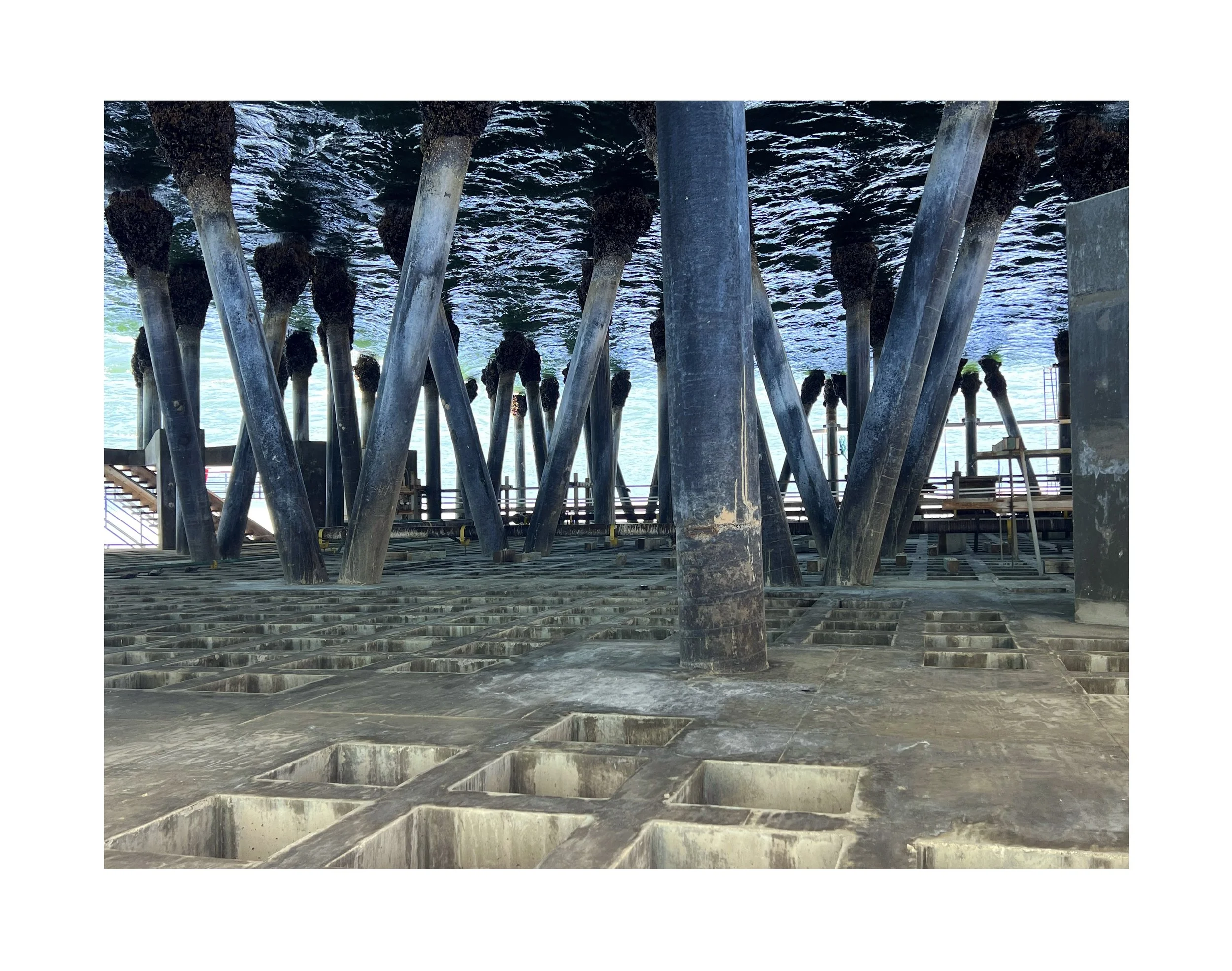 Structure en métal sous un pont ou une jetée, avec des piliers en croix et un sol en béton percé de nombreux orifices, vue de dessous.