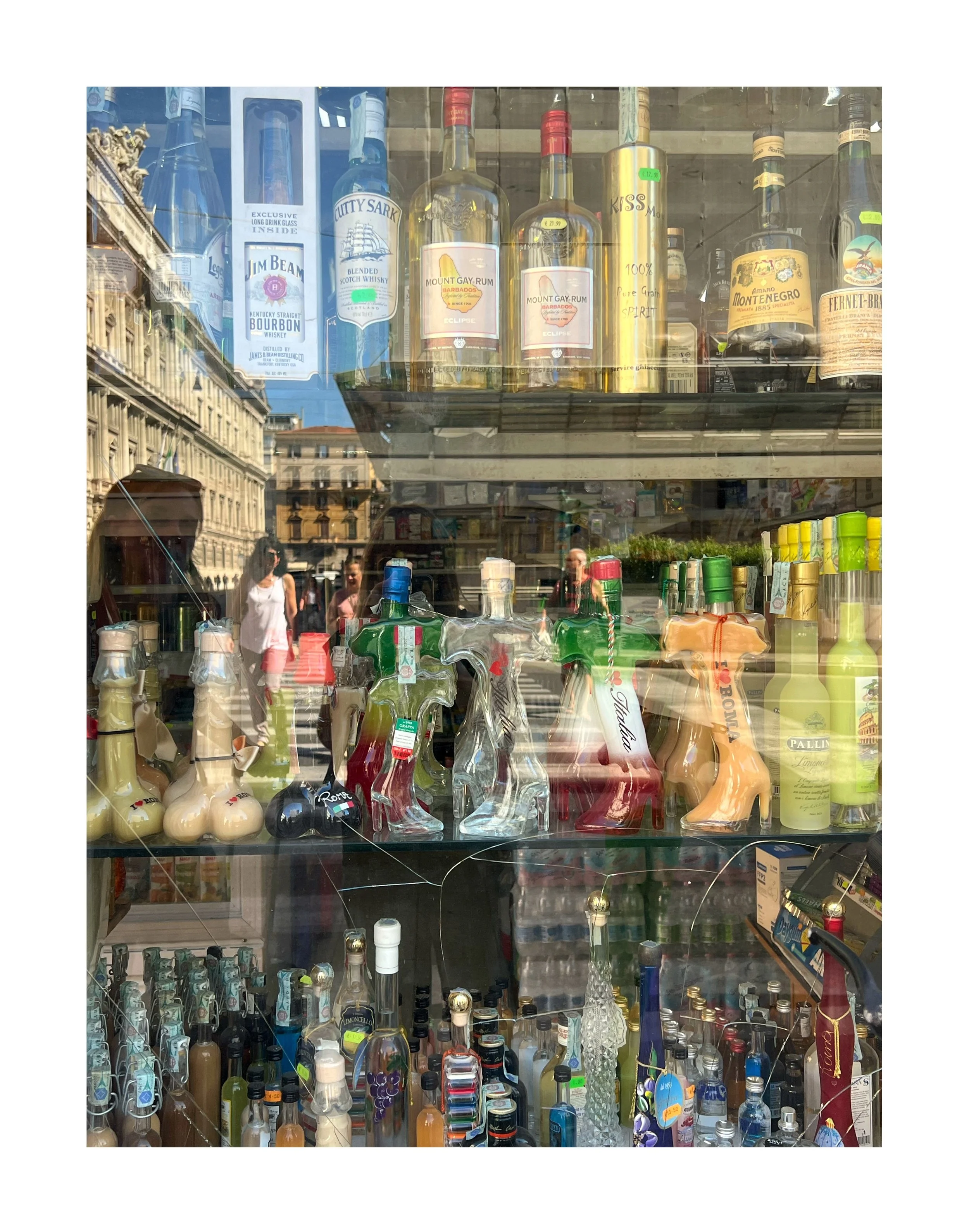 Vitrine d'une boutique avec des bouteilles d'alcool, des décorations en forme de chaussures et d'objets en verre, avec la réflexion de la rue et des passants visible à travers la vitre.