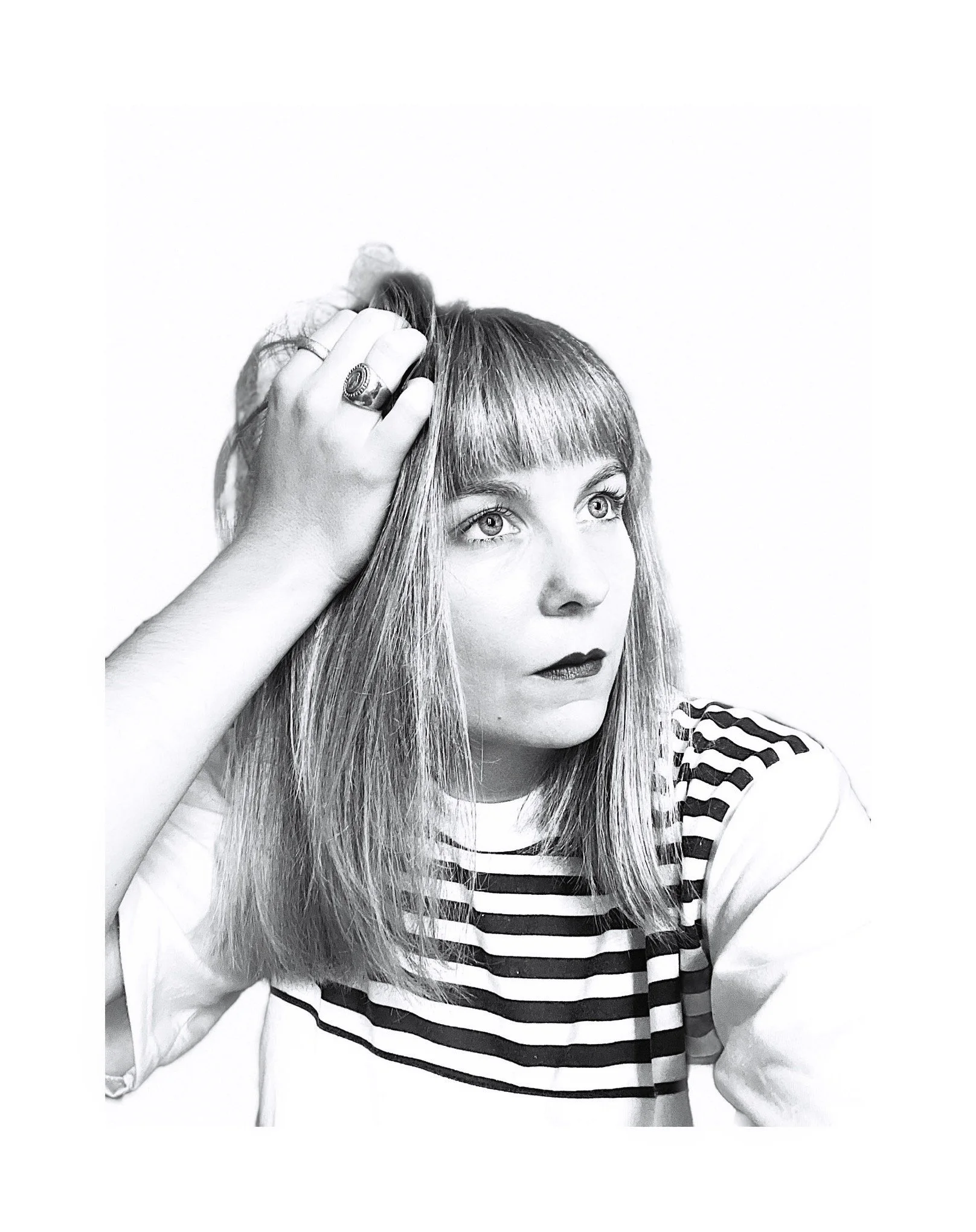 Une femme avec des cheveux longs et droits, portant un t-shirt à rayures, regarde vers la droite et touche ses cheveux avec sa main gauche.