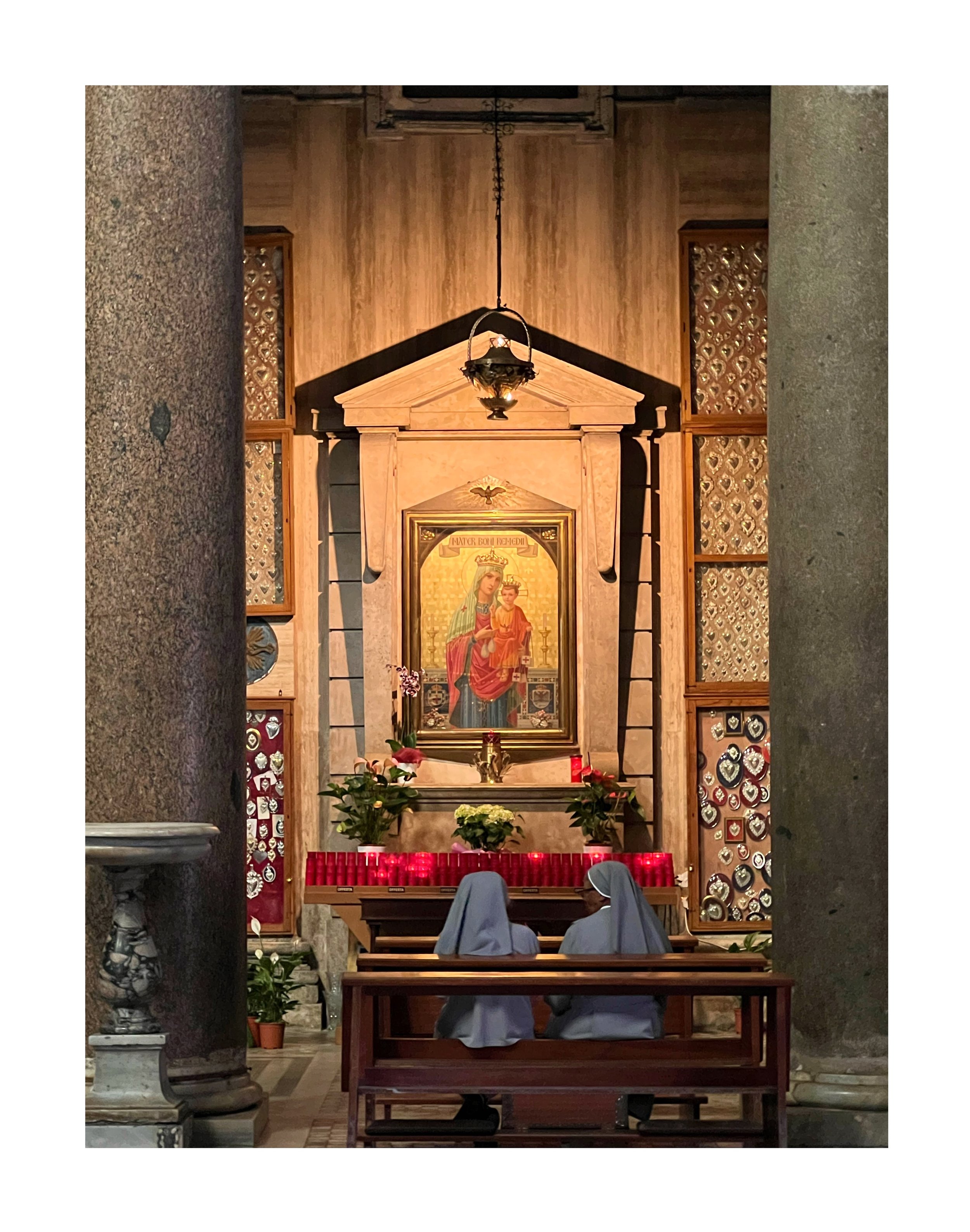 Deux religieuses prient dans une église devant une icône de la Vierge Marie et de l'enfant Jésus, avec des bougies rouges allumées et des fleurs autour.