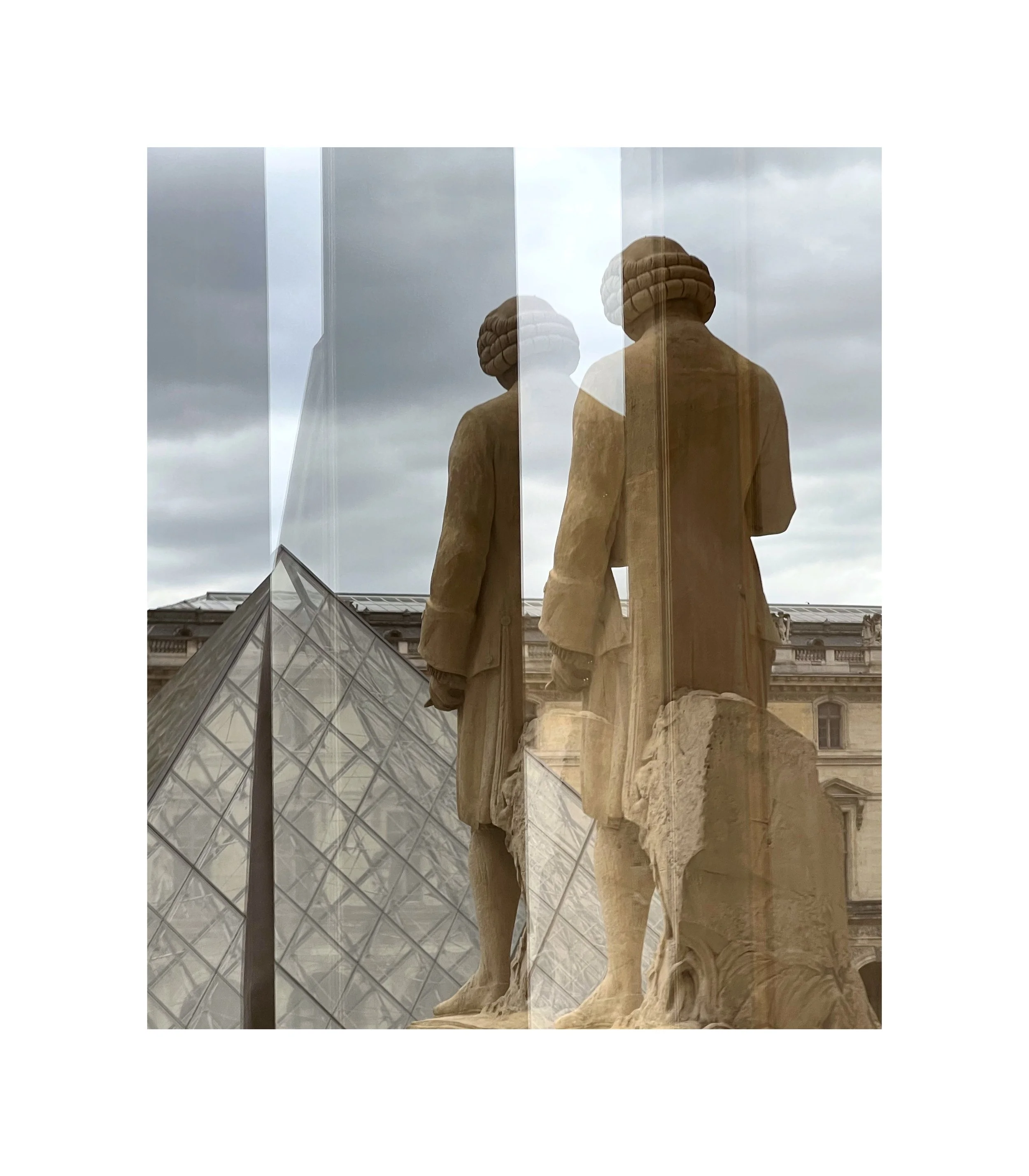 Vue de deux statues en pierre visibles à travers une vitre, avec le Louvre et le ciel nuageux en arrière-plan.