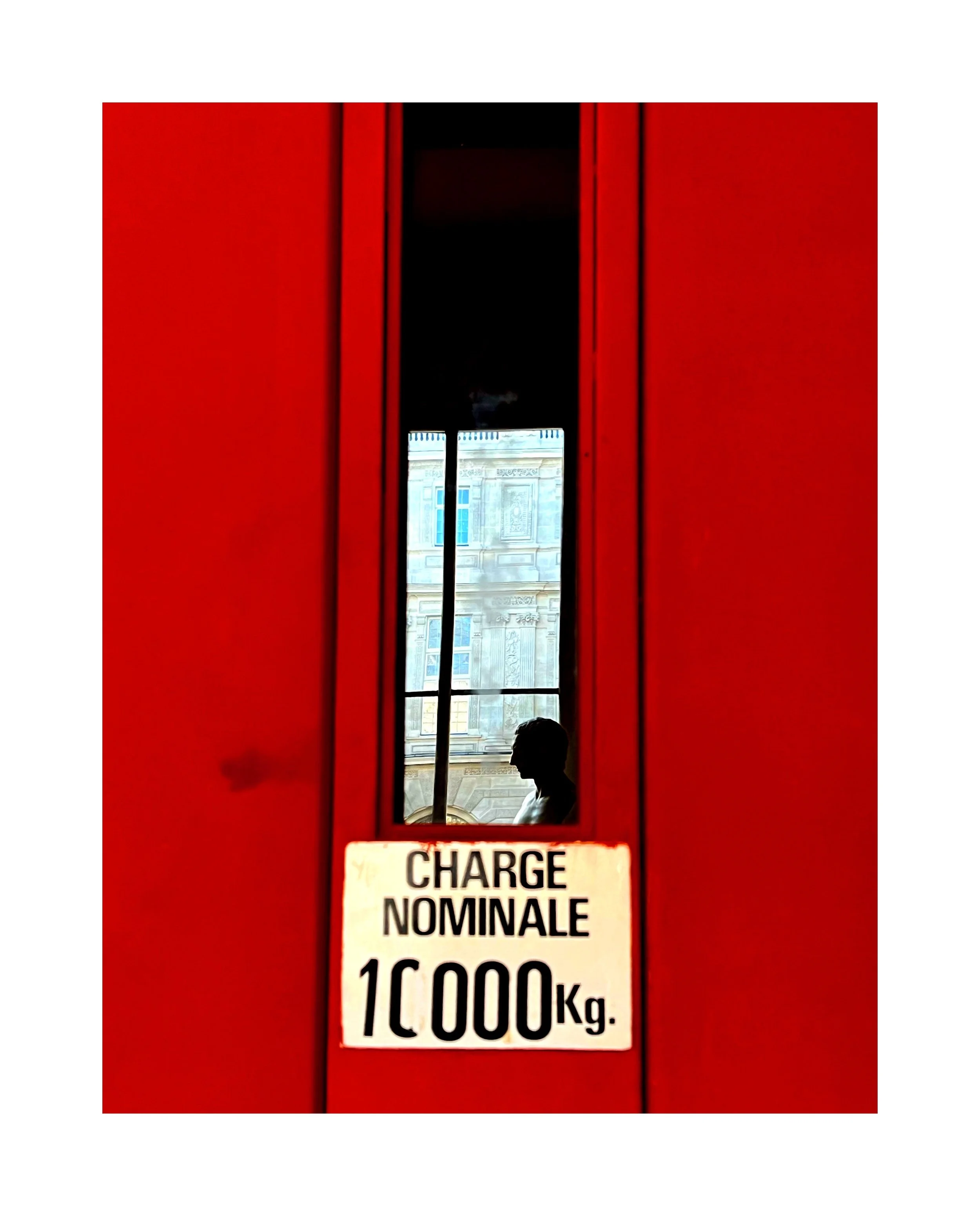 Une porte rouge avec une fenêtre en son centre, à travers laquelle on voit une silhouette d'homme et un bâtiment en arrière-plan. Sur la porte, il y a une pancarte indiquant "Charge Nominale 1000 Kg".