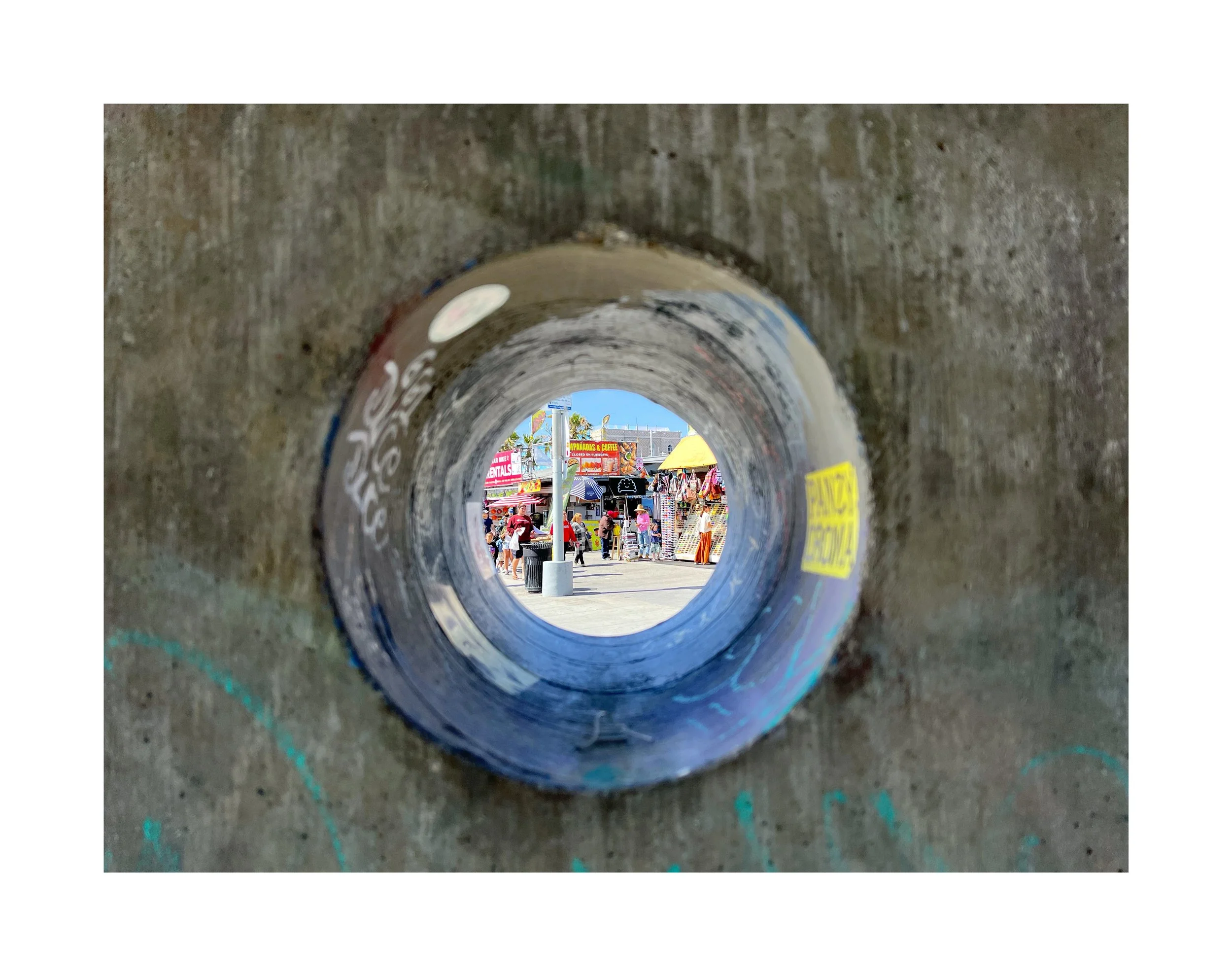 Vue d'un festival ou marché à travers un trou dans un mur en béton, avec stands, personnes, parasols colorés et un ciel en arrière-plan.