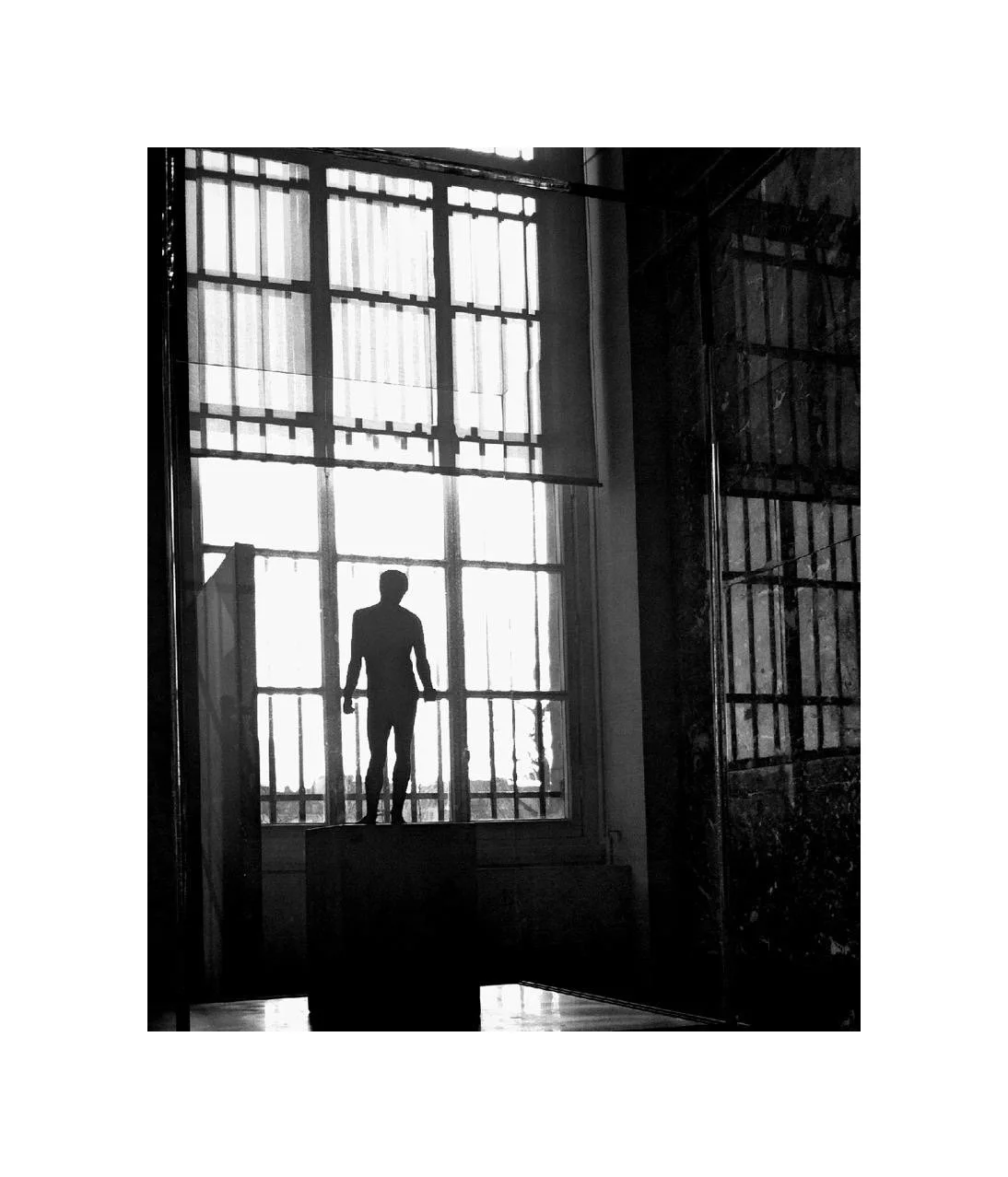 Silhouette d'un homme debout devant une grande fenêtre avec une grille, dans une pièce sombre.