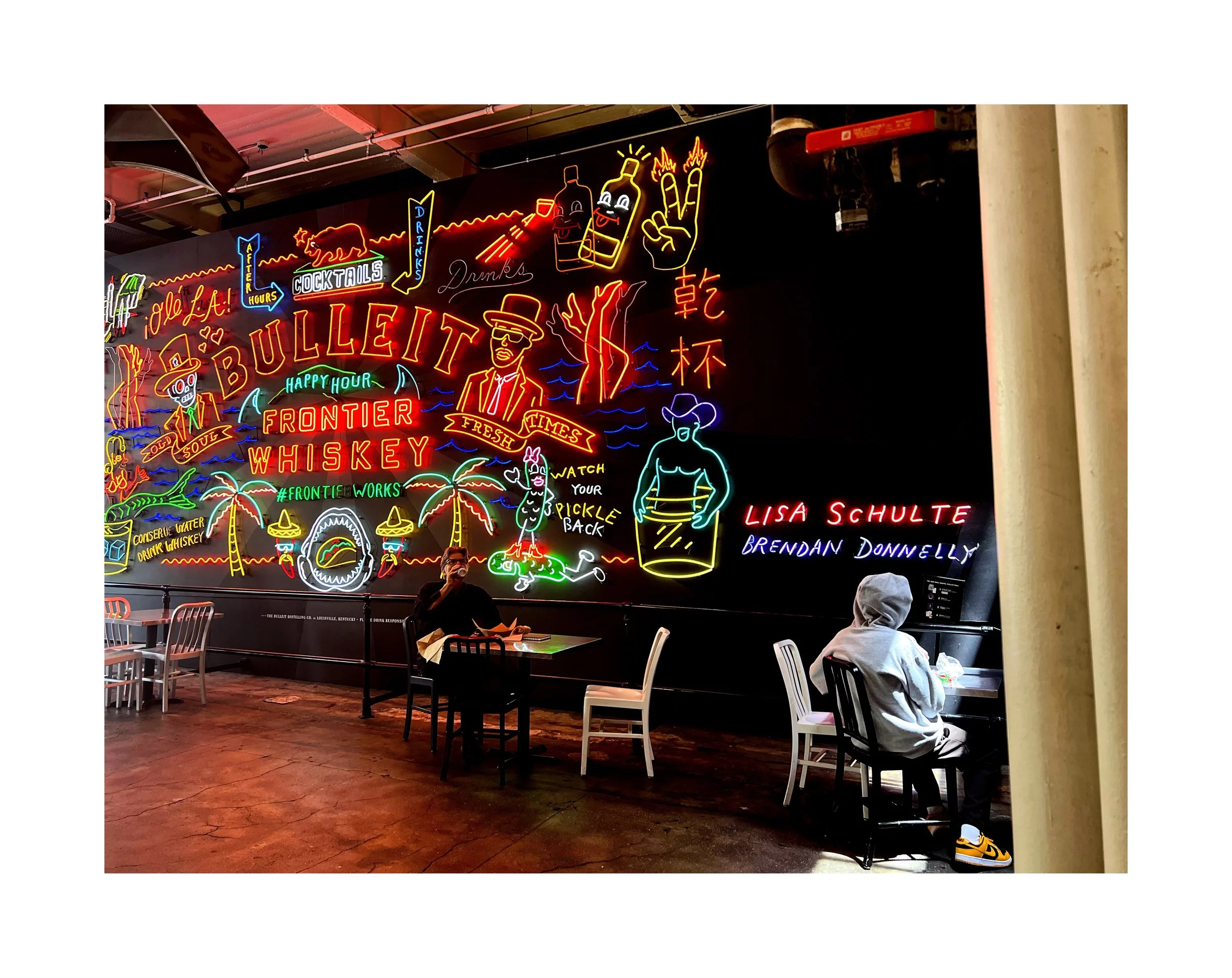 Mur de néons colorés avec des illustrations de bouteilles, verre, palmier, homme portant un chapeau, personnage avec boucles d'oreilles, et texte comme « Bullitt », « Whiskey », « Frontier », « Lisa Schulte », et « Brendan Donnelly » dans un restaura