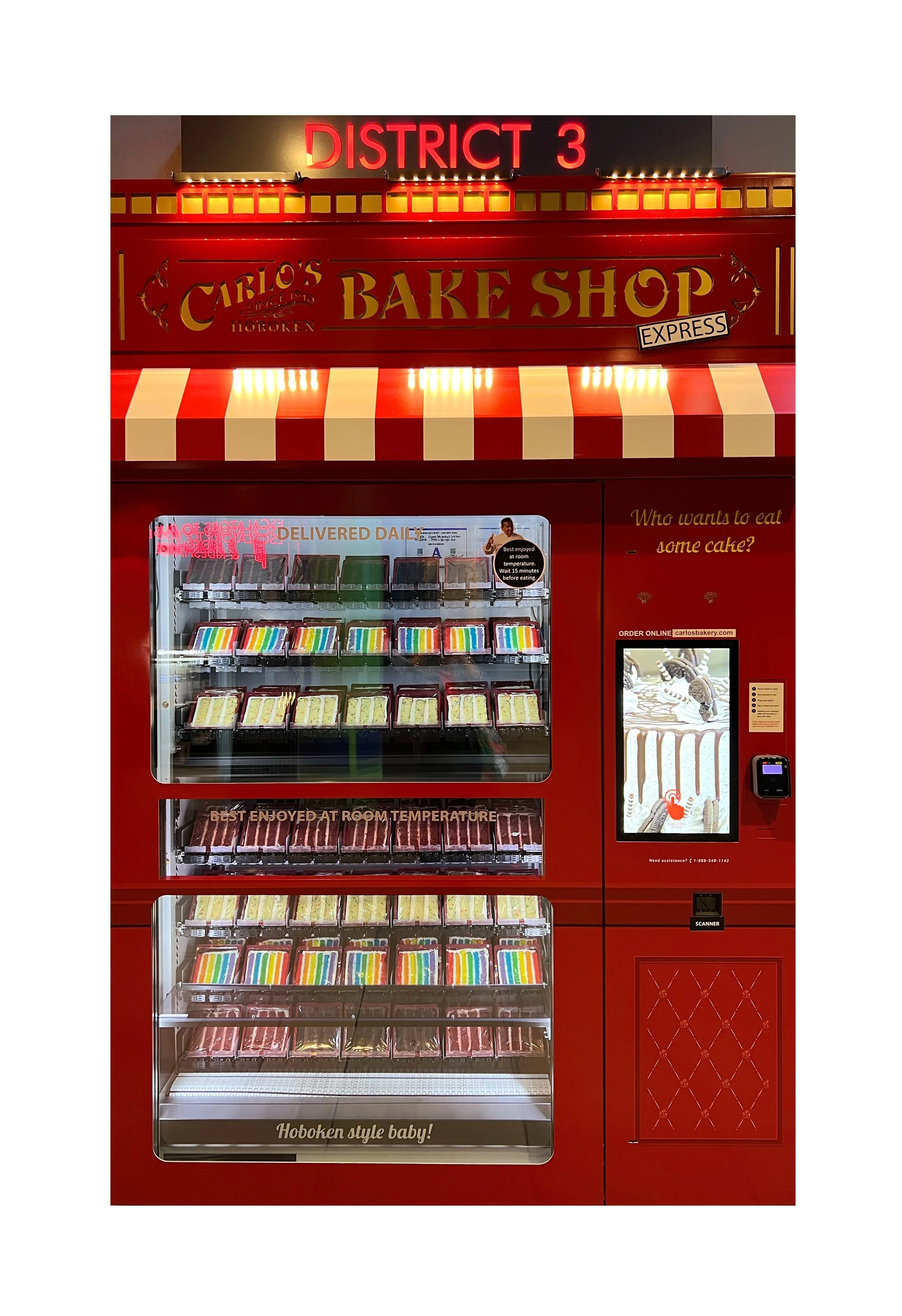 Vending machine à gâteaux avec le panneau 'Carlos Bake Shop' et le texte 'District 3' en haut. La machine propose différents gâteaux et desserts avec une caisse automatique à droite.