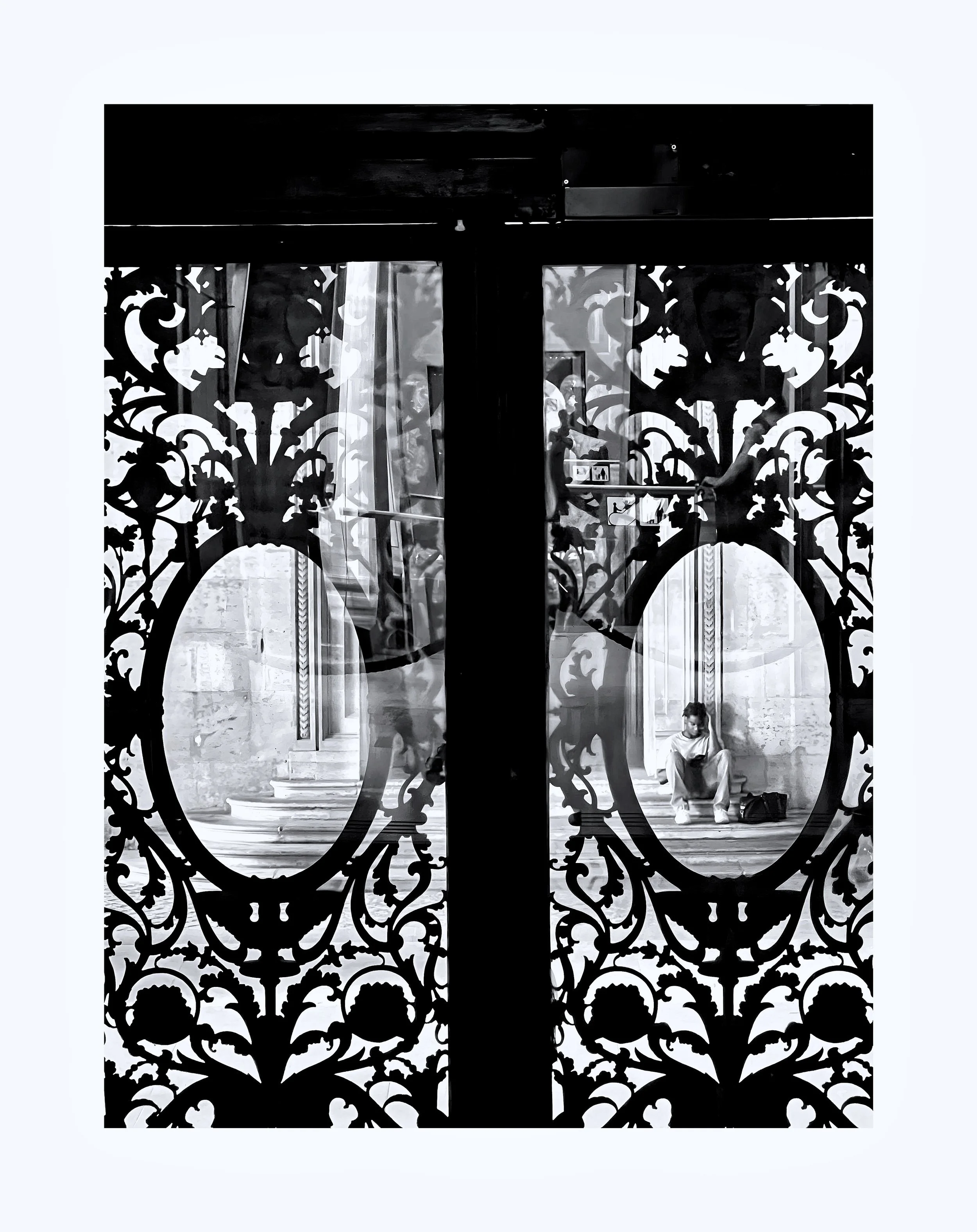 Une personne assise sur les marches d'un bâtiment, vue à travers une porte en fer forgé ornée de motifs floraux, en noir et blanc.