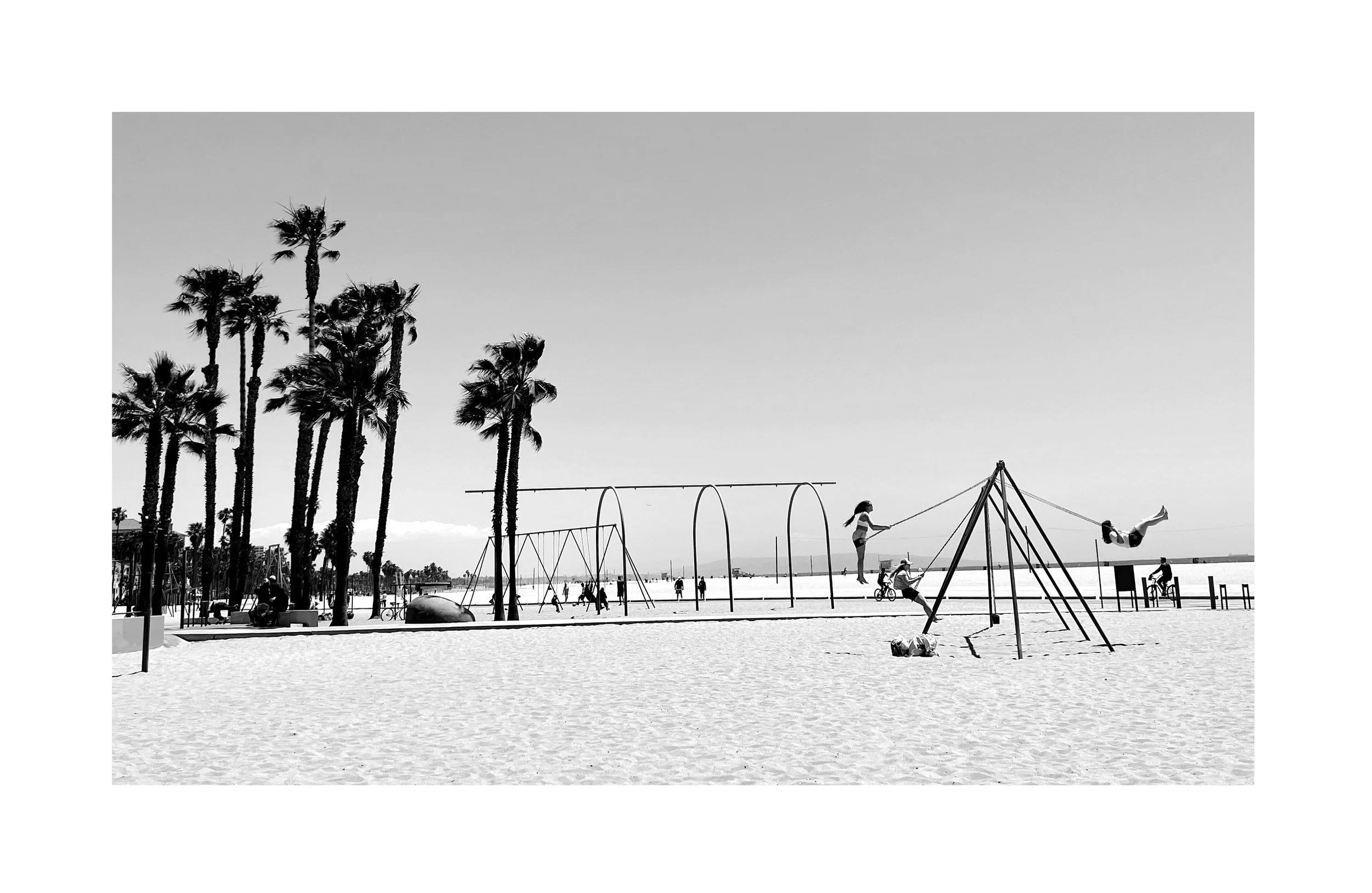 Plage avec des palmiers, des enfants jouant sur une balançoire, une aire de jeux en métal et des personnes en arrière-plan, en noir et blanc.