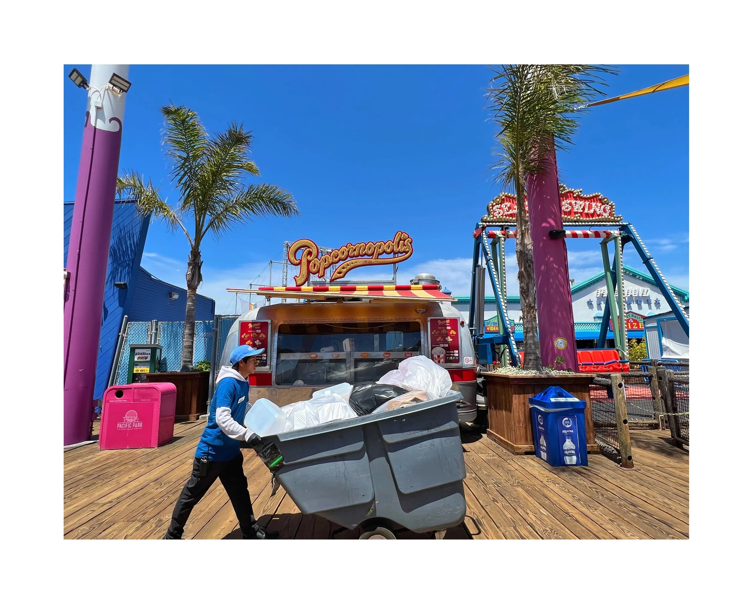 Une personne en uniforme pousse une chariot rempli de déchets dans un parc d'attractions avec des attractions comme une grande roue et une maison hantée, sous un ciel bleu avec des palmiers.