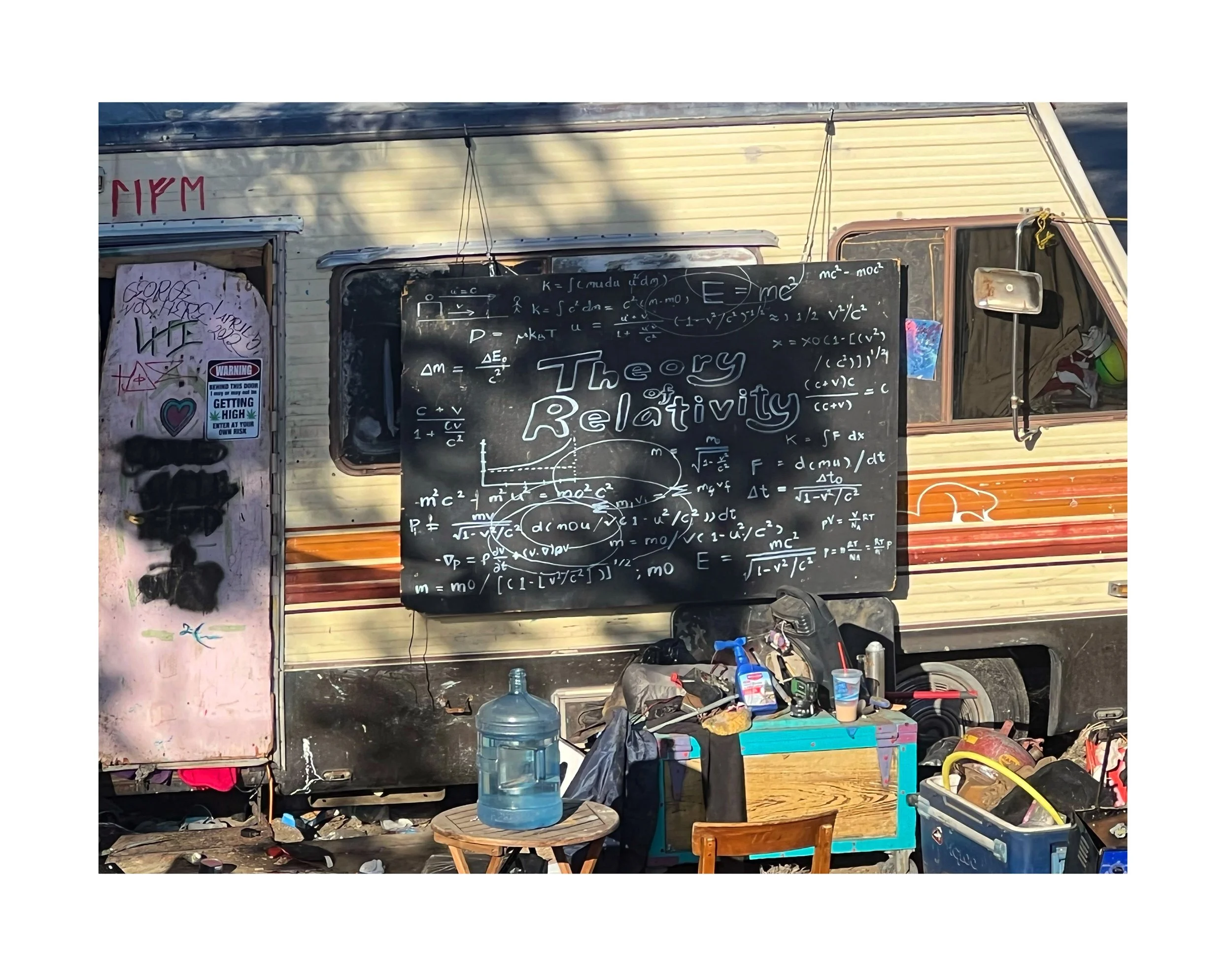 Une caravane avec un tableau noir affichant des équations physiques et le titre 'Theory of Relativity' écrit en blanc. Devant, il y a un petit meuble en bois avec une cruche d'eau, et plusieurs objets comme un spray, un gobelet, et des outils dispers