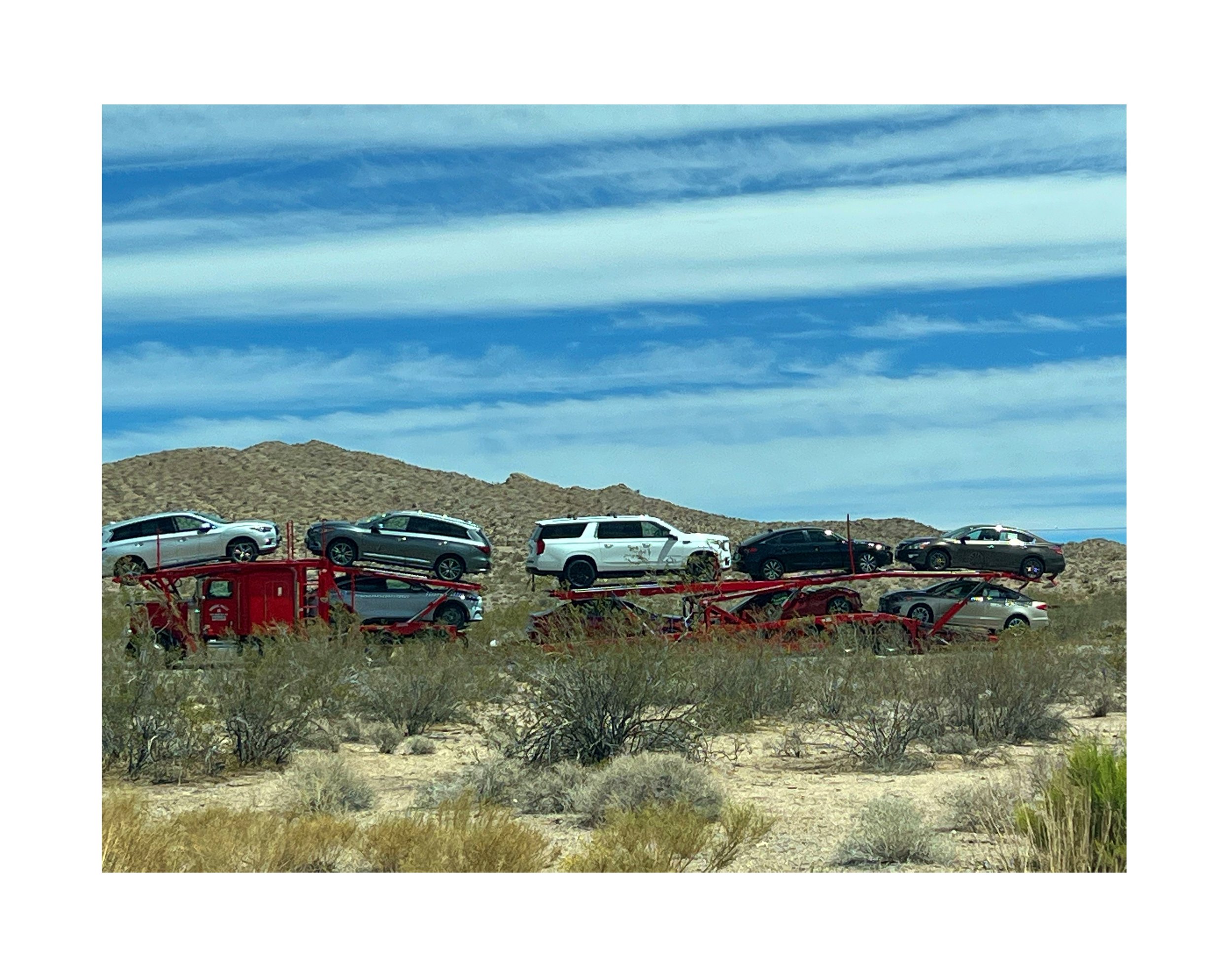 Une caravane de voitures sur un bus escalier rouge dans un désert avec des buissons et un ciel bleu avec des nuages.