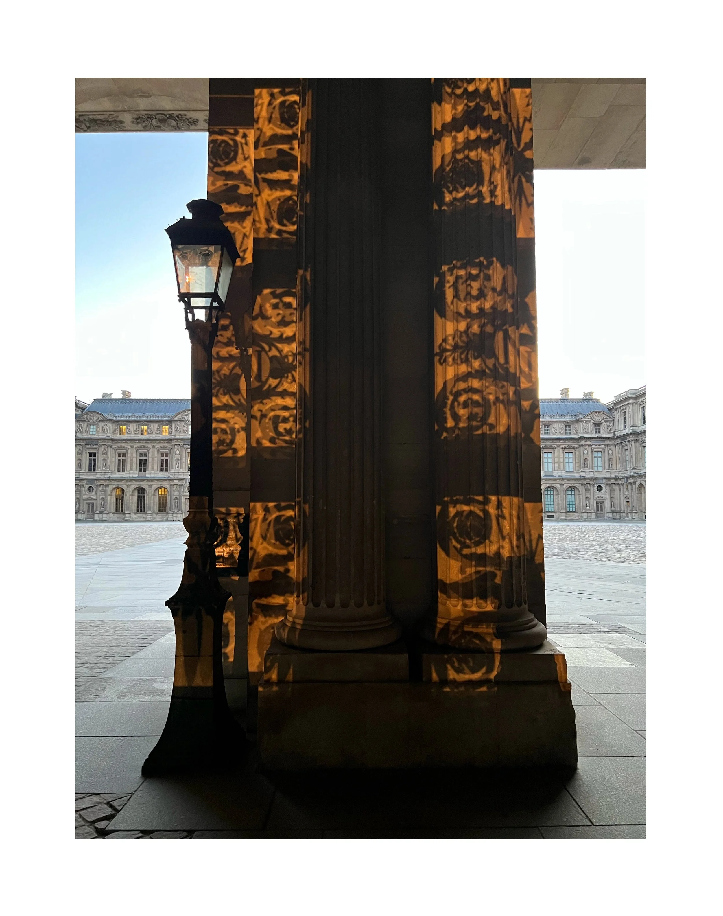 Reflet d'une lampadaire et de colonnes classiques dans une surface réfléchissante, avec un bâtiment architectural en arrière-plan, ciel clair.