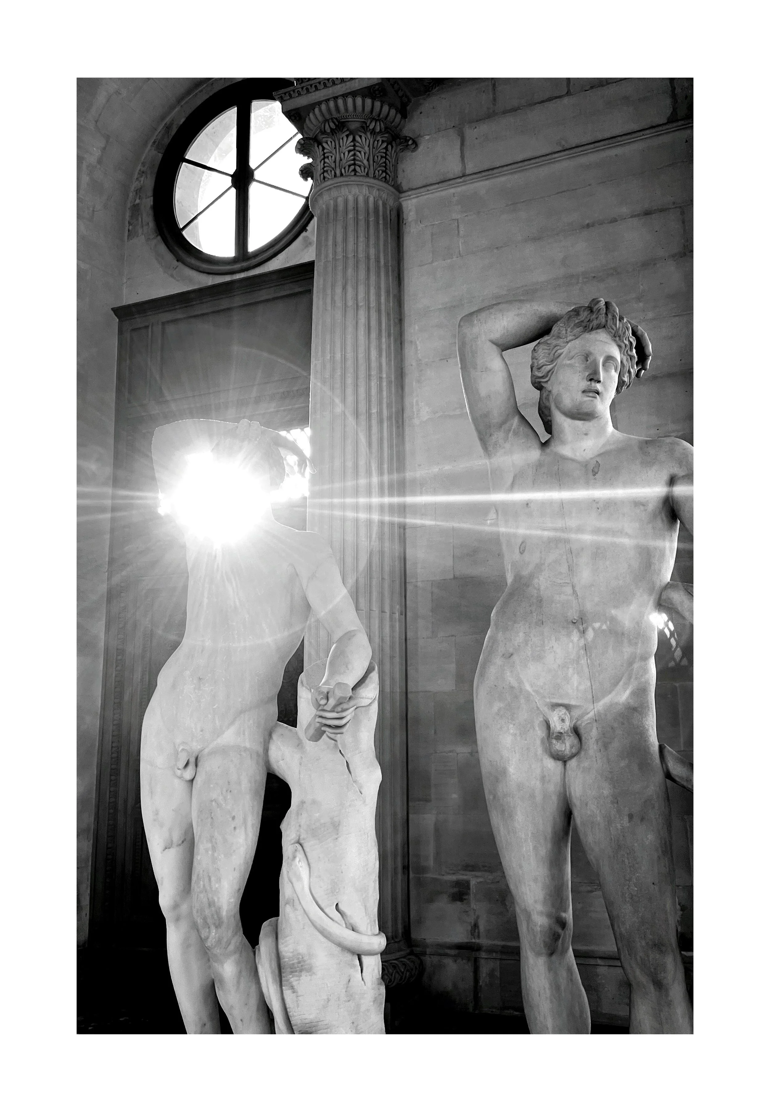 Photographie en noir et blanc de deux sculptures masculines classiques, dont une avec une main derrière la tête. La lumière du soleil brille à travers une figure, créant un éclat puissant. Il y a une colonne ornementée et une fenêtre ronde en arrière