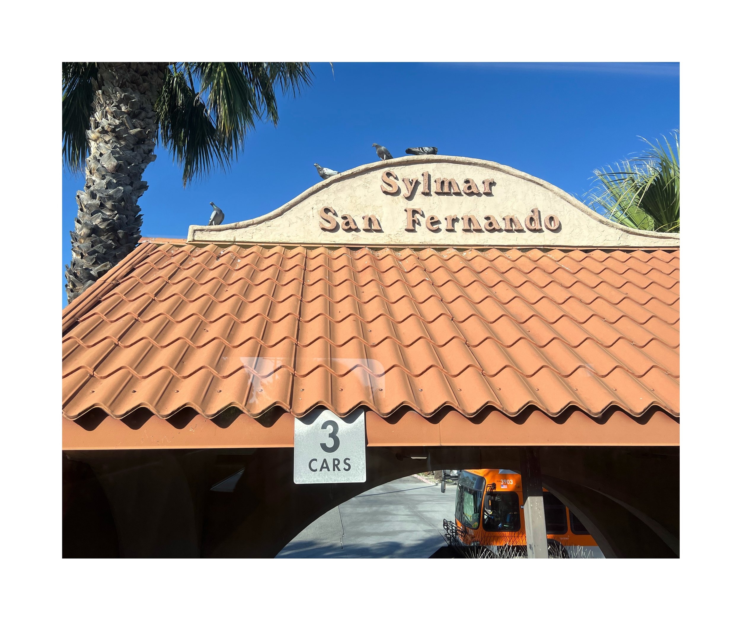 Entrée du parking de Sylmar San Fernando avec un toit en tuiles rouges et un panneau indiquant '3 CARS'. Sur le toit, il y a plusieurs pigeons et des palmiers en arrière-plan contre un ciel bleu clair.
