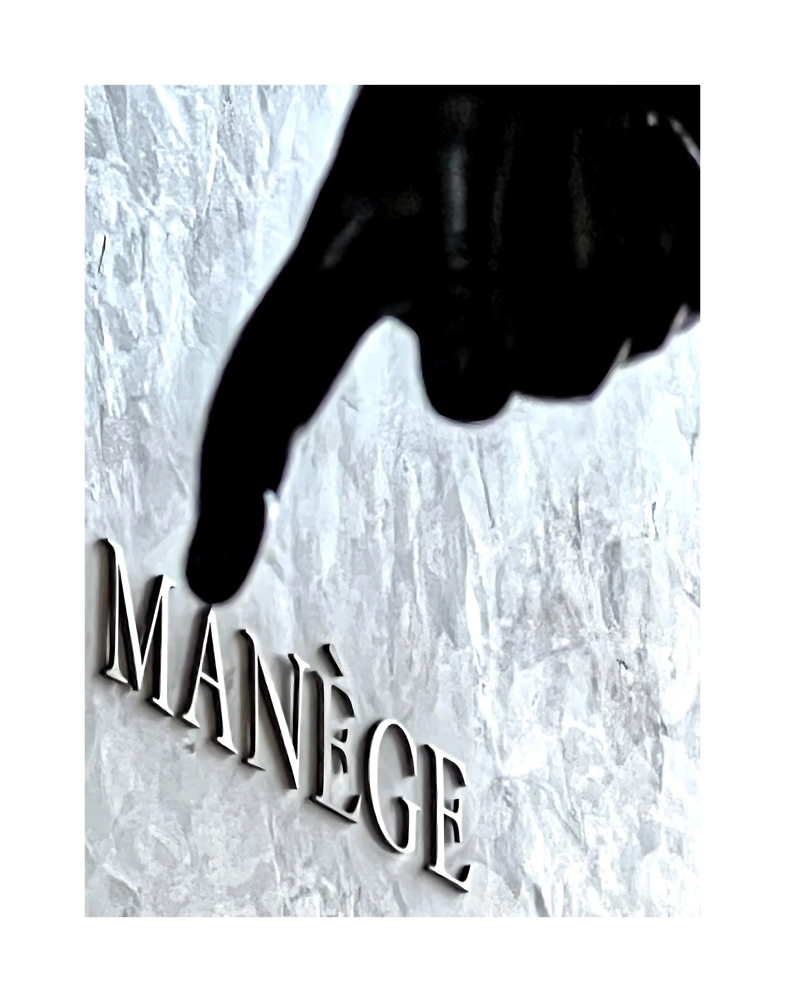 Une main en plastique noir pointant vers le nom « MANÉGE » écru en relief sur un mur en pierre.