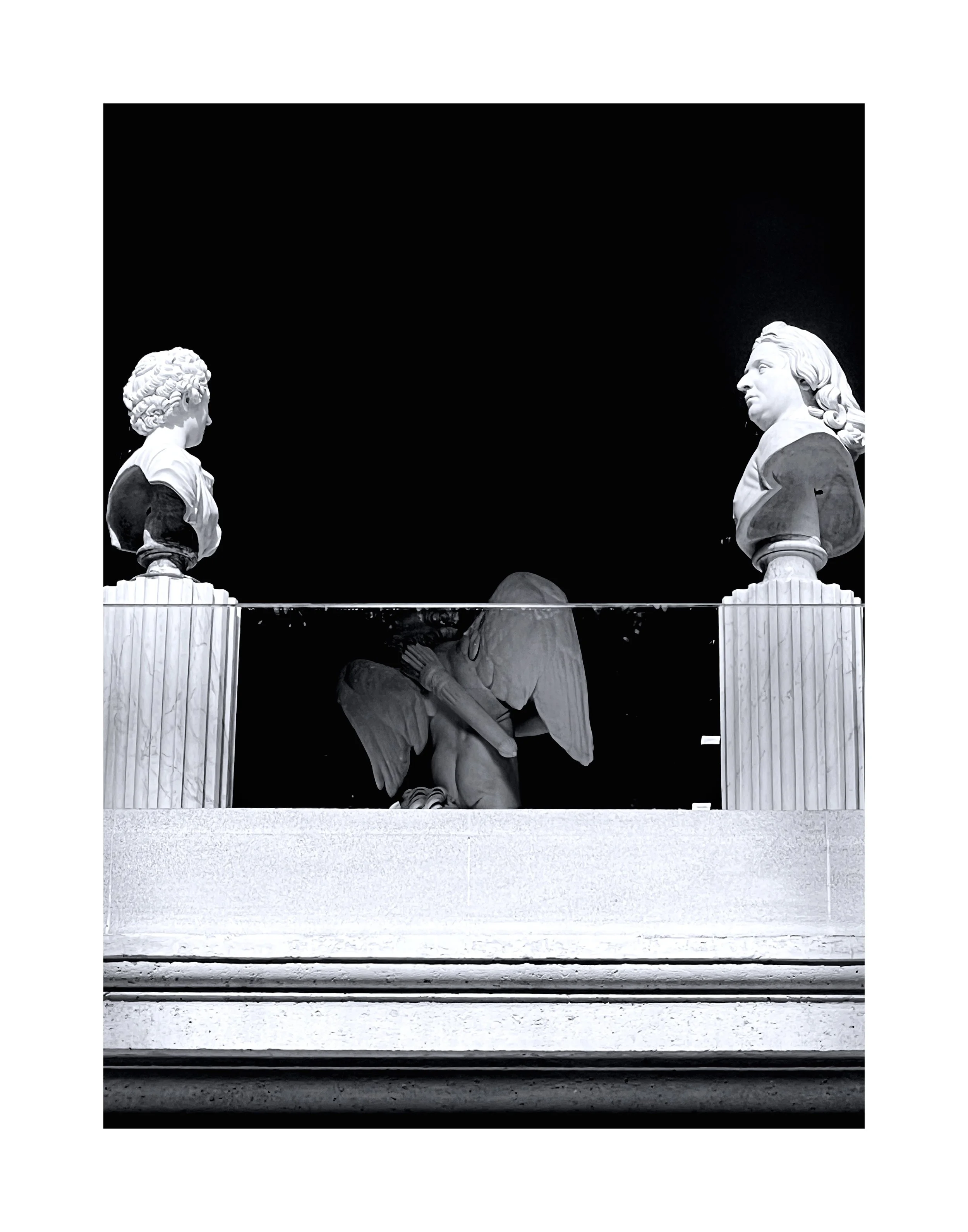 Sculptures classiques en marbre exposées sur un socle, dont deux bustes sur piédestaux et une sculpture d'ange. La scène est en noir et blanc.