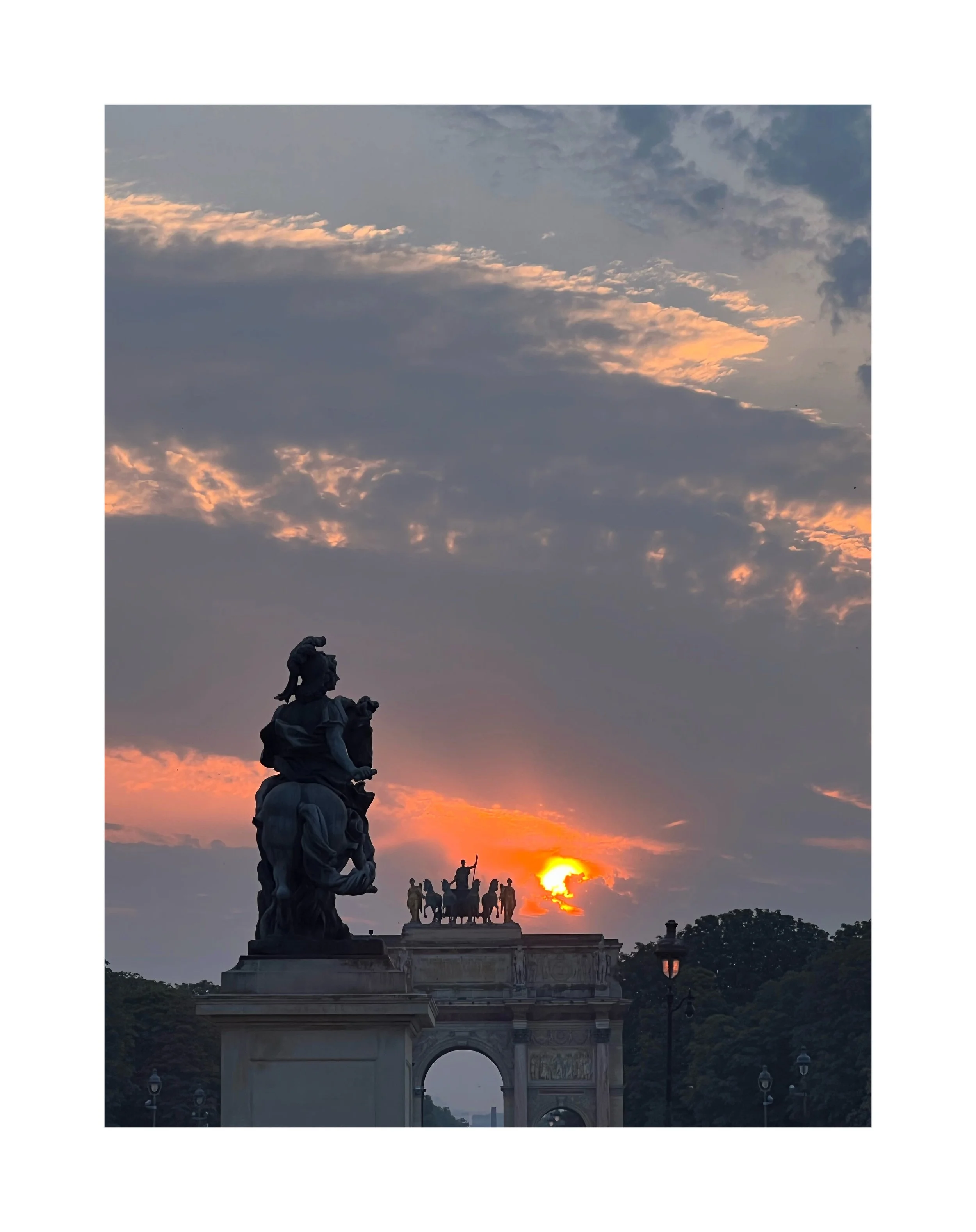 Statues sur une fontaine ou un monument, avec le coucher de soleil en arrière-plan et un ciel partiellement nuageux.