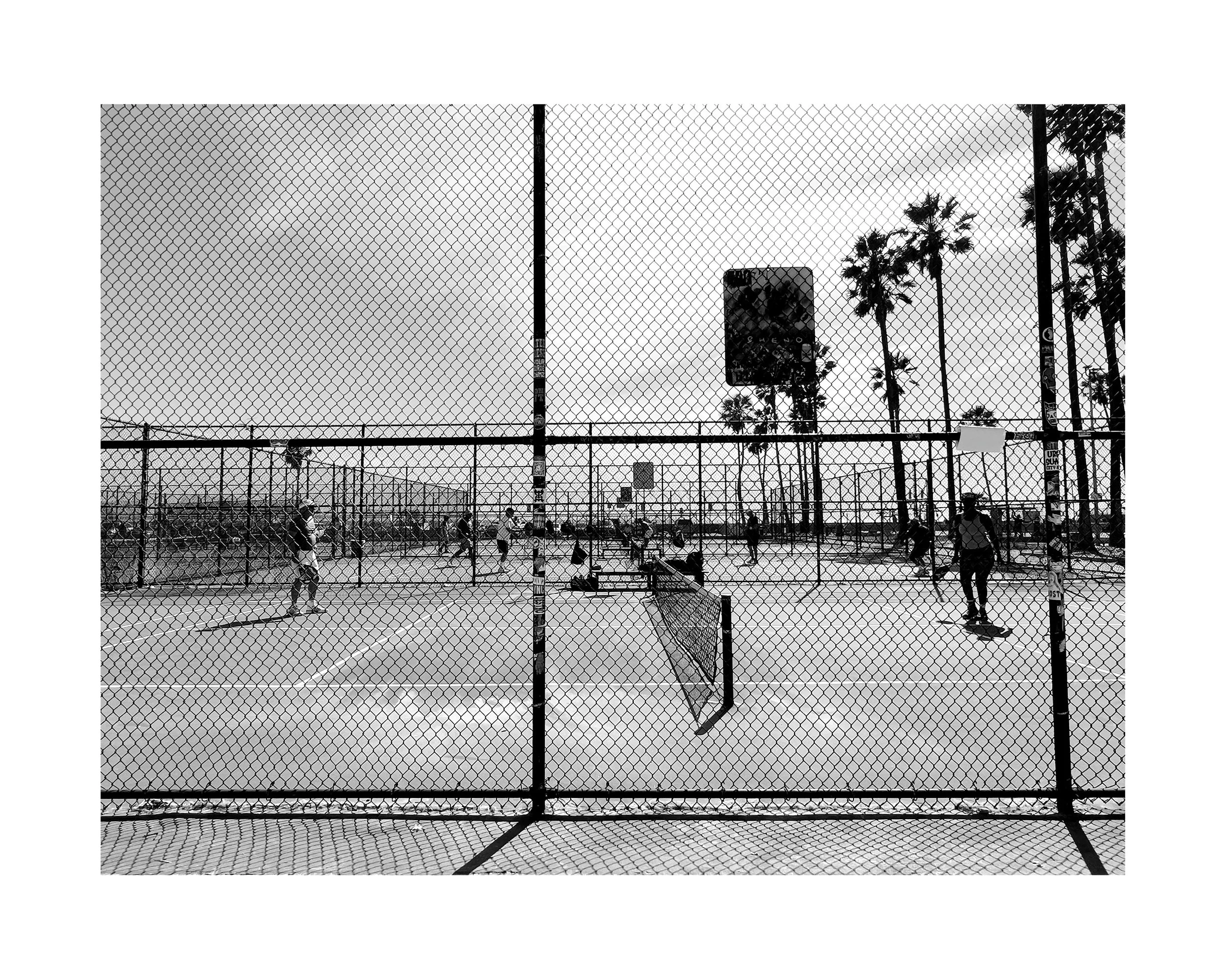 Un terrain de basket-ball extérieur vu à travers une clôture en métal, avec plusieurs joueurs en activité, et des palmiers en arrière-plan