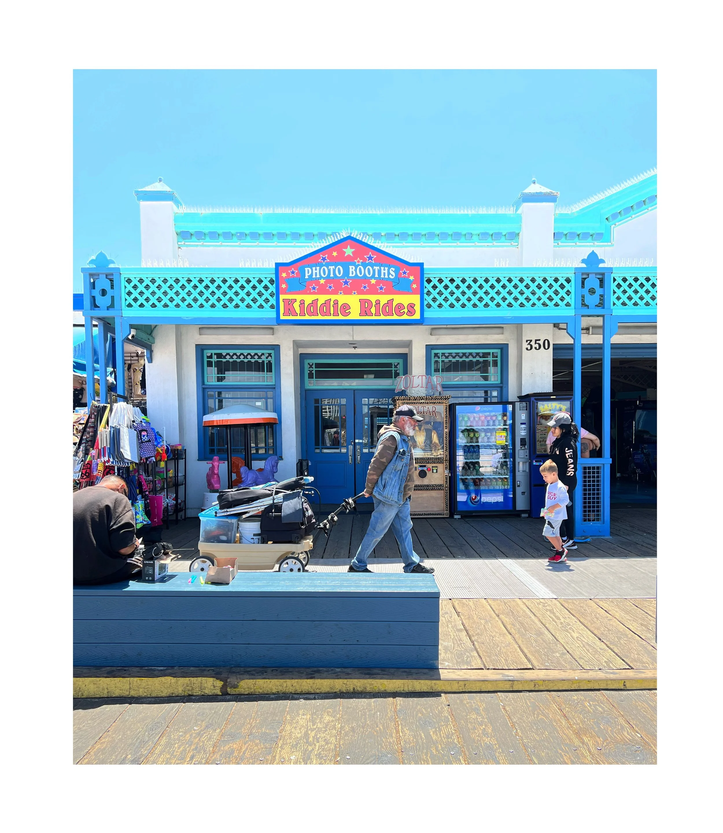 Façade d'un kiosque de photo avec panneau 'Kiddie Rides', situé au bord de la mer sous un ciel bleu, avec des personnes qui passent, dont un homme tirant un chariot et une famille regardant.