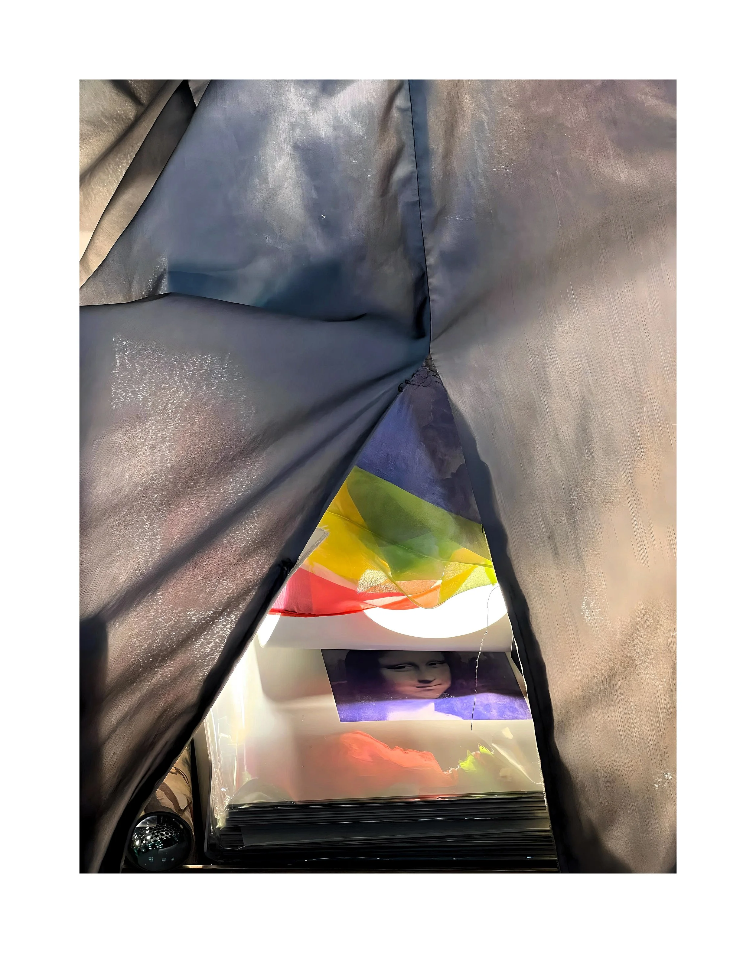 Une collection de photographies ou d'œuvres d'art dans un album, vues à travers une ouverture dans une couverture ou un tissu coloré, avec une silhouette humaine ou un visage féminin visible sur une des images.