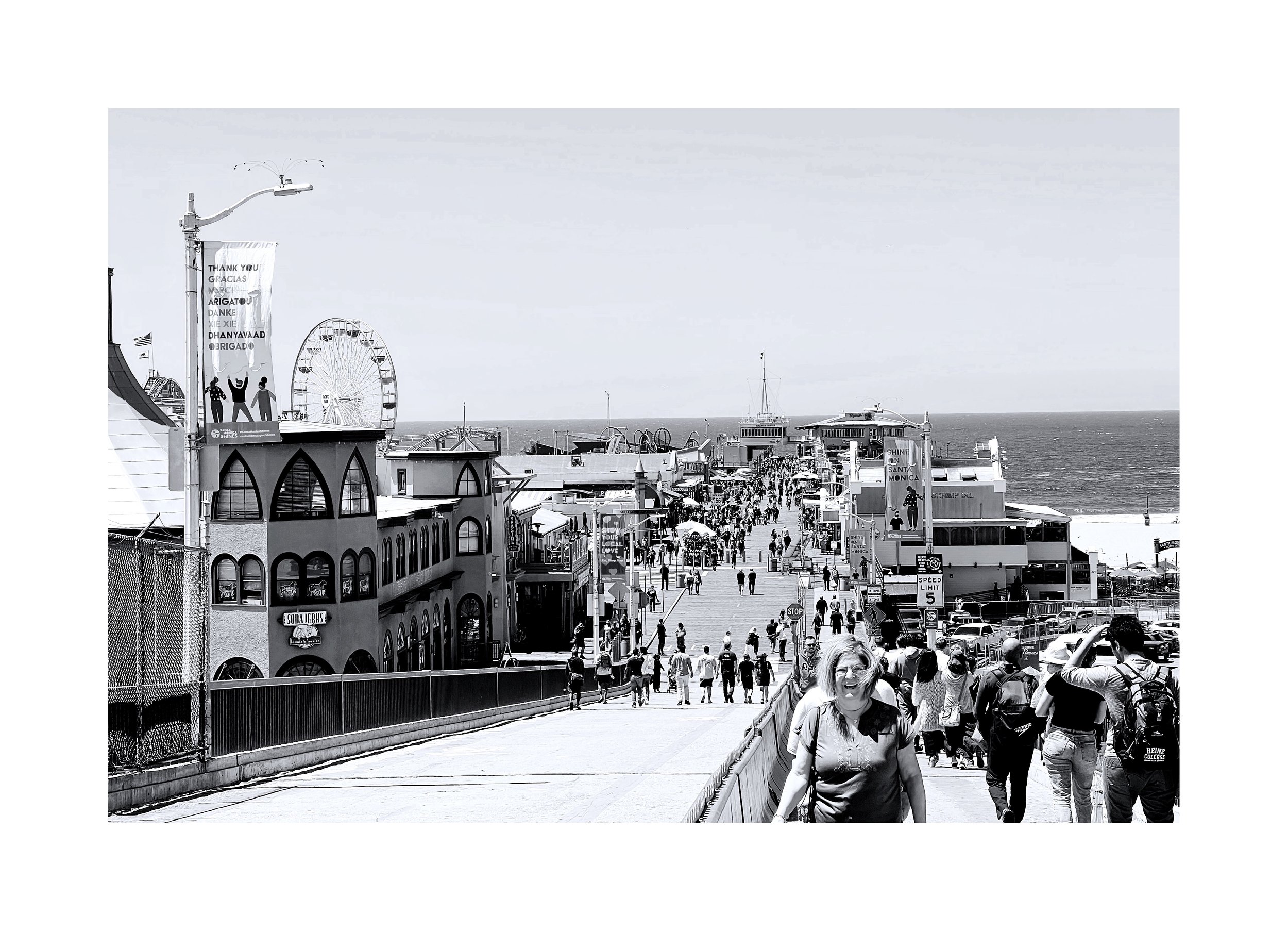 Une scène de promenade en bord de mer avec de nombreuses personnes qui marchent. On aperçoit des bâtiments colorés, une grande roue, une grande illusion d'une fête avec des attractions de style carnaval, et l'océan en arrière-plan.