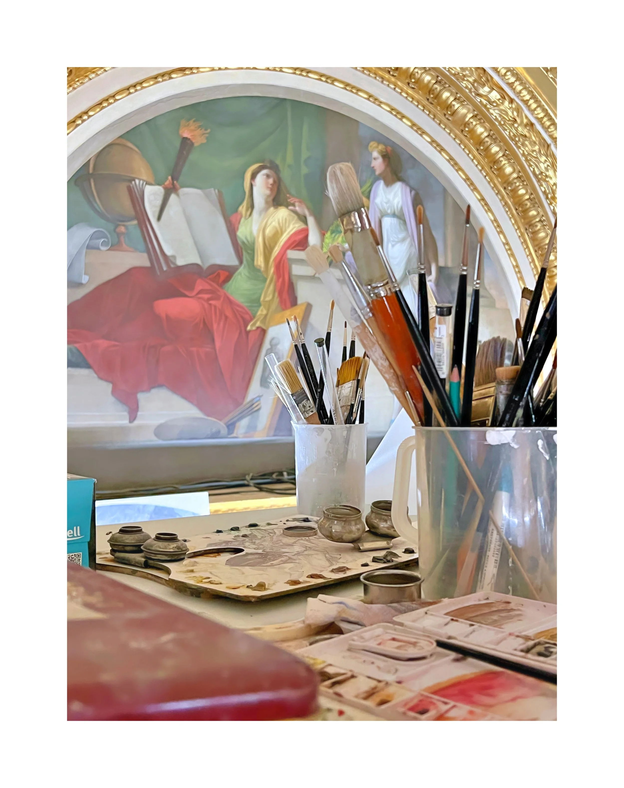 Établi d'artiste avec des pinceaux dans des contenants en plastique, une palette de peinture, et un tableau mural représentant une scène classique avec deux femmes, une assise avec un livre et une autre debout, dans un décor ornemental doré.