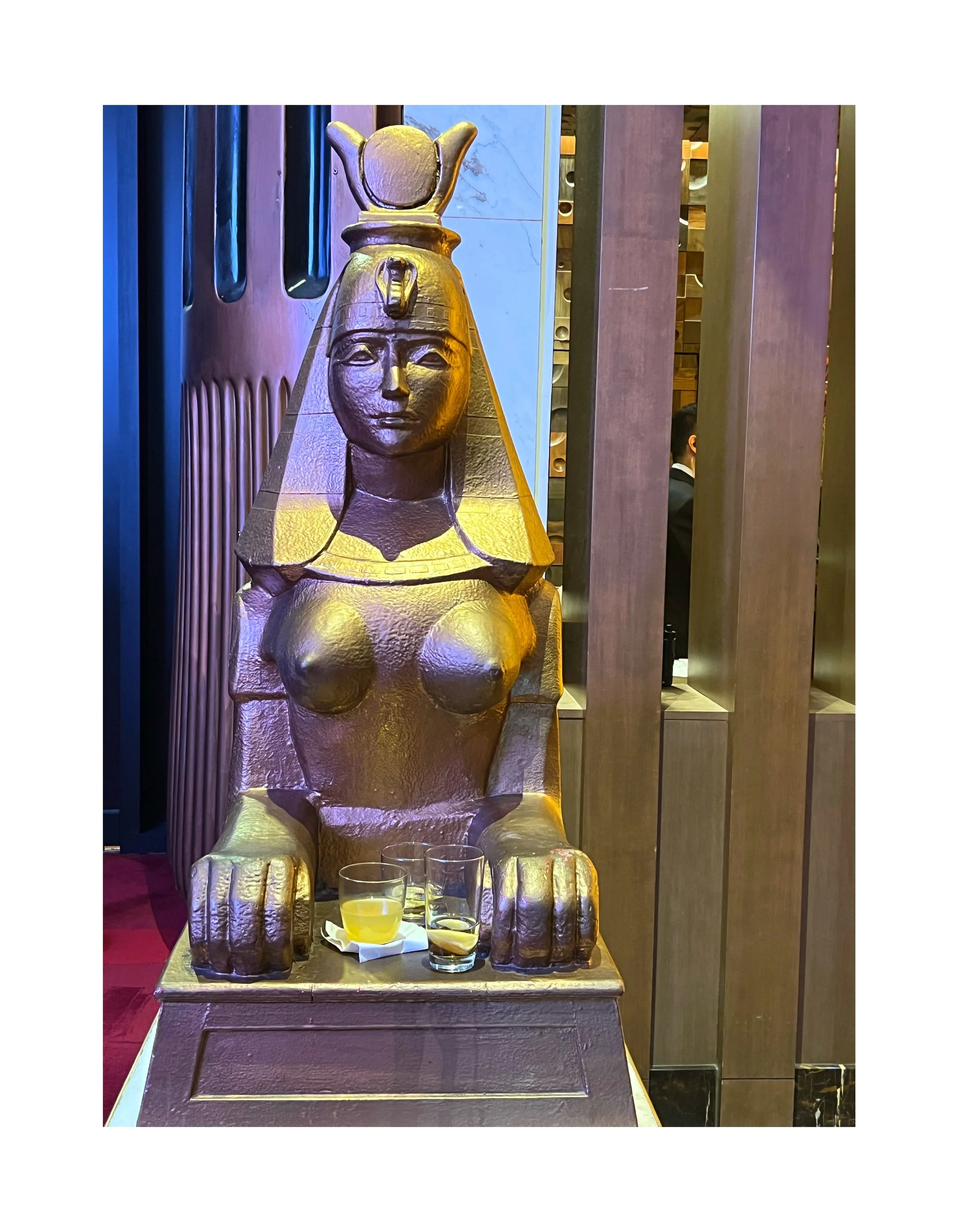 Statue en bois représentant une figure égyptienne, avec une tête de pharaon et un corps féminin, située dans un restaurant avec des boissons en face.