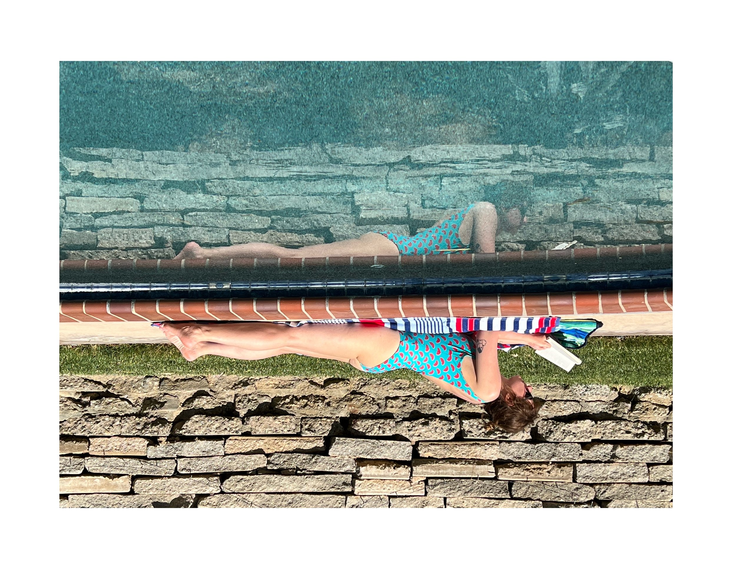 Femme allongée près d'une piscine, portant un maillot de bain coloré, lisant un livre, avec un mur de pierre en arrière-plan.
