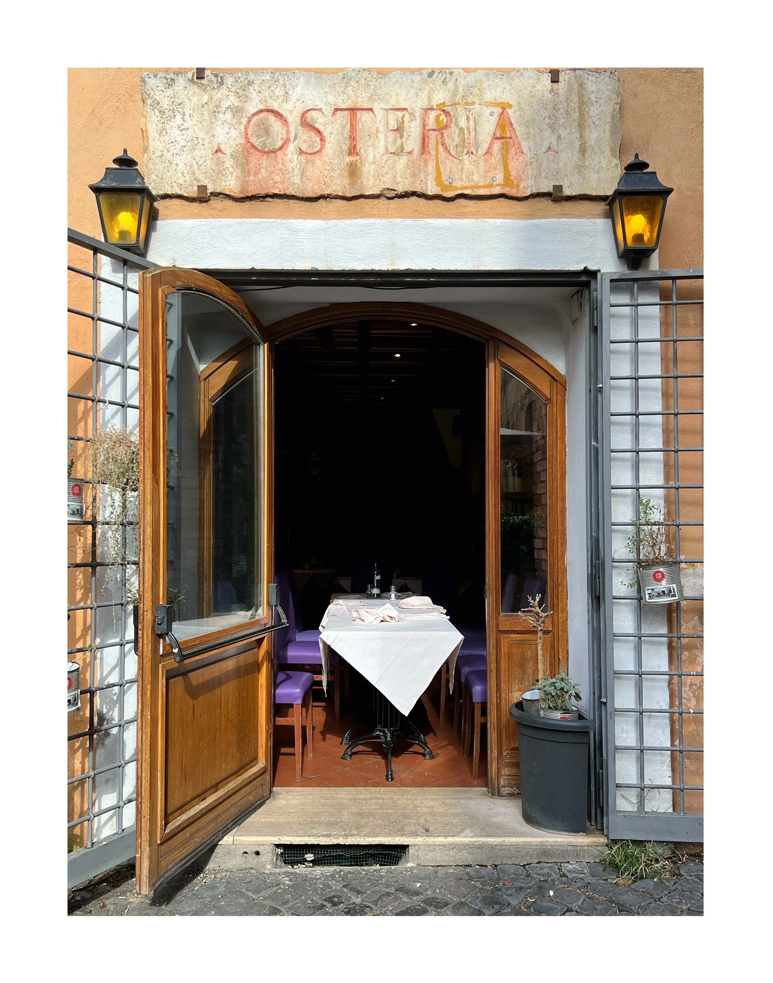 Vue de l'entrée d'une Osteria avec portes en bois ouvertes, tables à manger à l'intérieur avec nappe blanche, deux lanternes noires à l'extérieur, et un panneau indiquant le nom de l'établissement, décoration rustique avec plantes en pot.