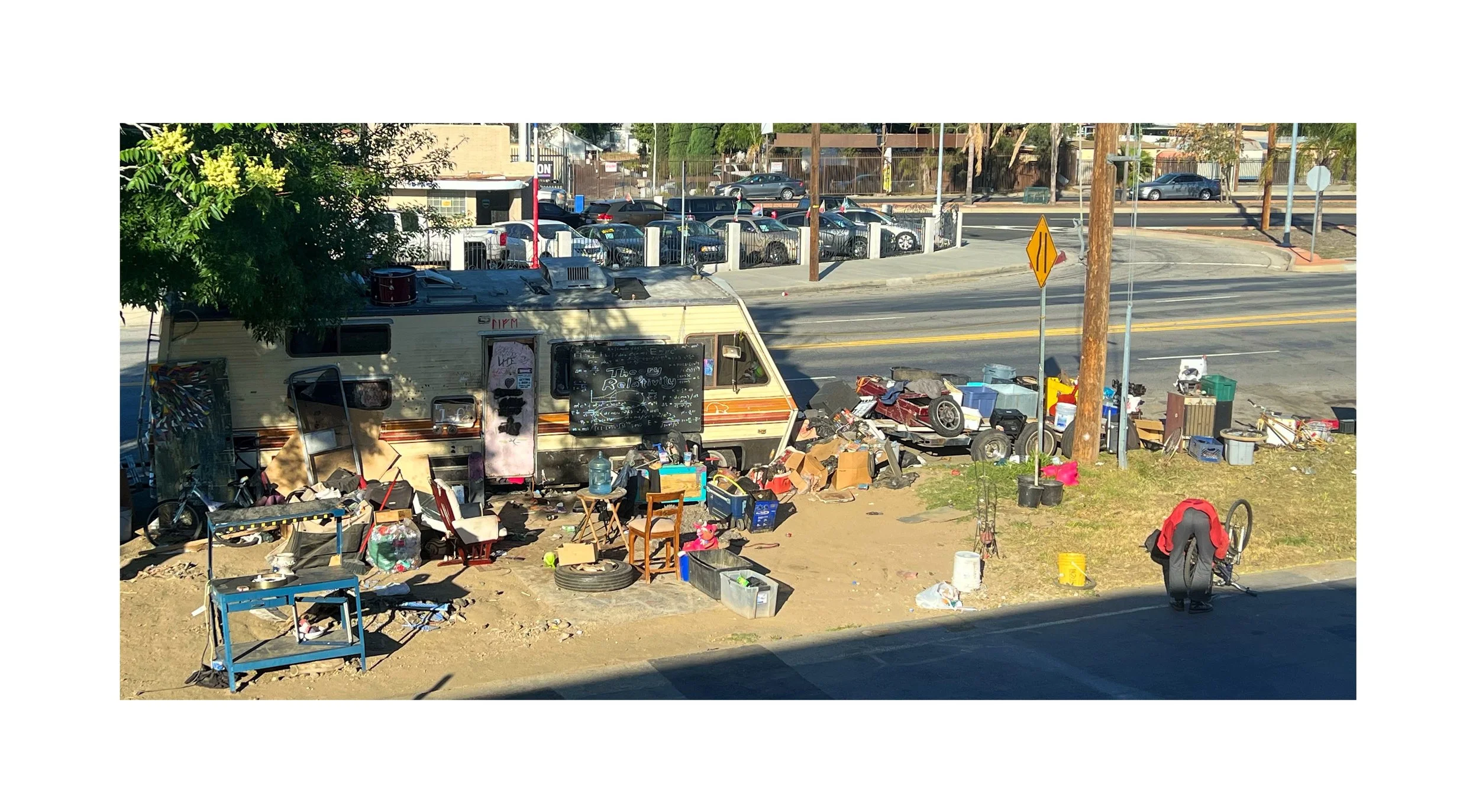 Vente de biens d'occasion en plein air avec un campeur à l'arrière-plan, des objets éparpillés, y compris des meubles, des boîtes, et un vélo, près d'une rue ensoleillée.