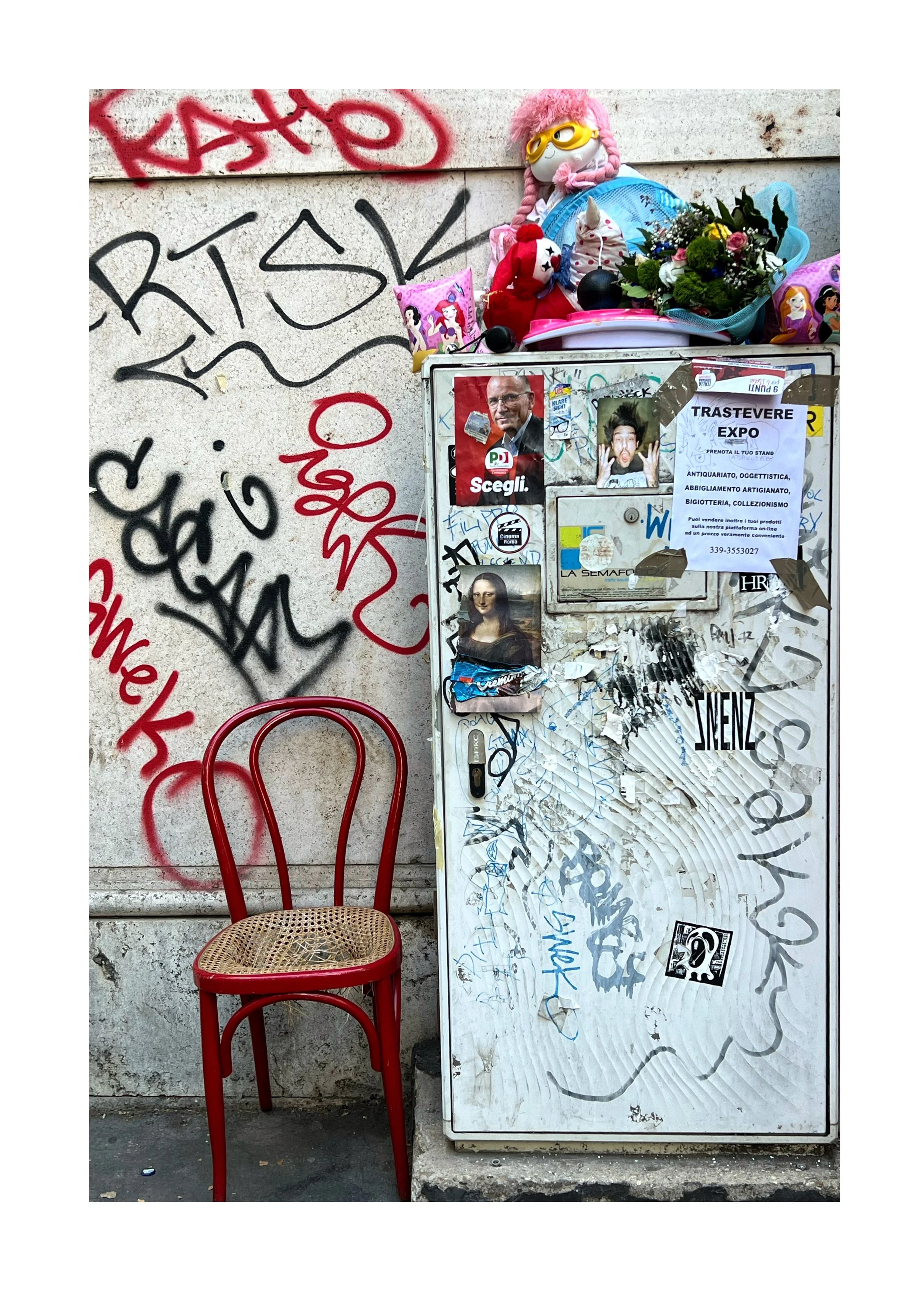 Une armoire à gauche d'une chaise rouge en bois avec un siège en rotin, devant un mur graffité. Sur le dessus de l'armoire, il y a un bouquet de fleurs, des peluches, et des coussins avec des personnages de dessins animés, notamment de Disney.