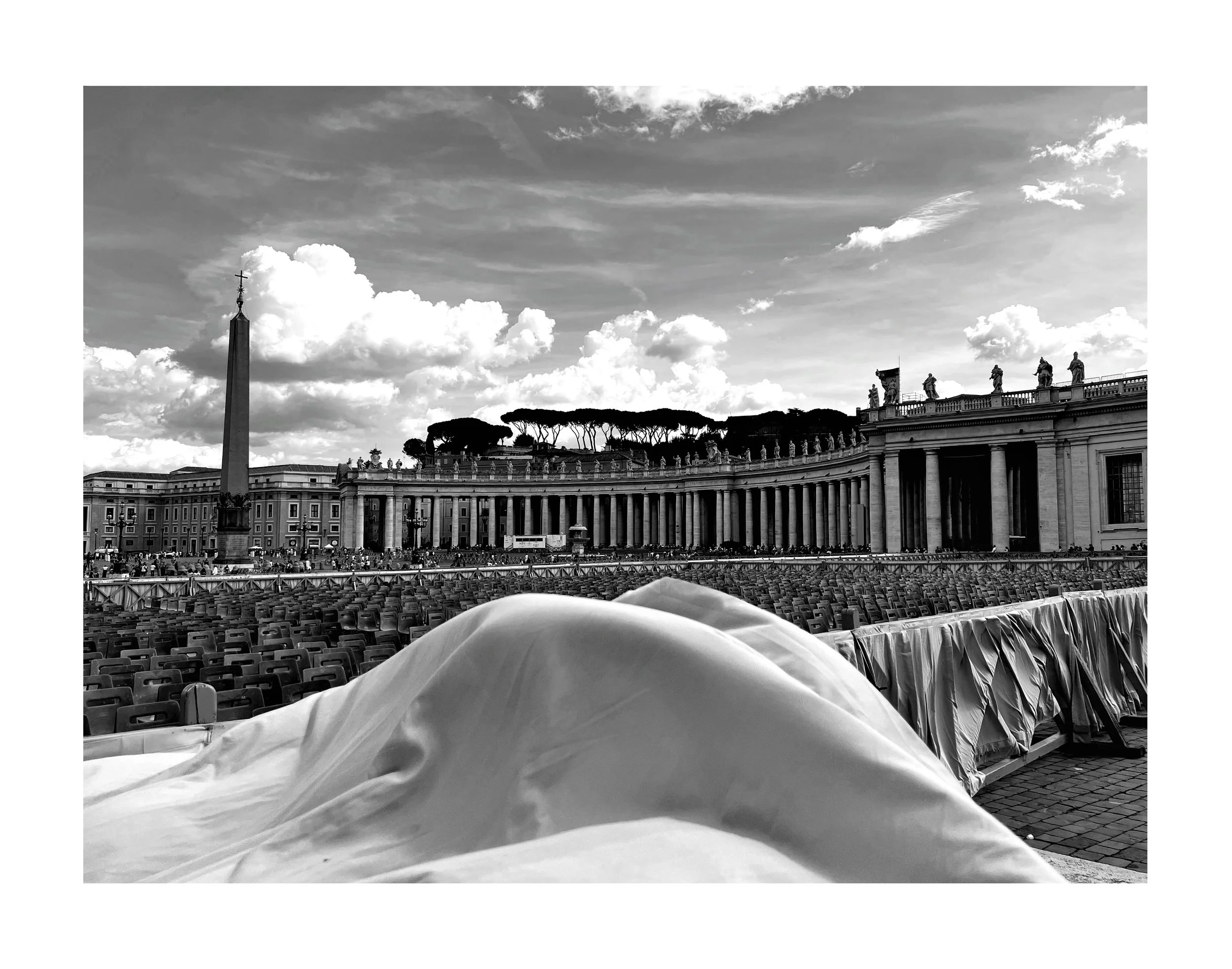 Vue en noir et blanc de la Place Saint-Pierre au Vatican, avec un drap ou un tissu placé en premier plan, des chaises alignées, une colonne avec une croix, et le Palais Apostolique en arrière-plan sous un ciel nuageux.