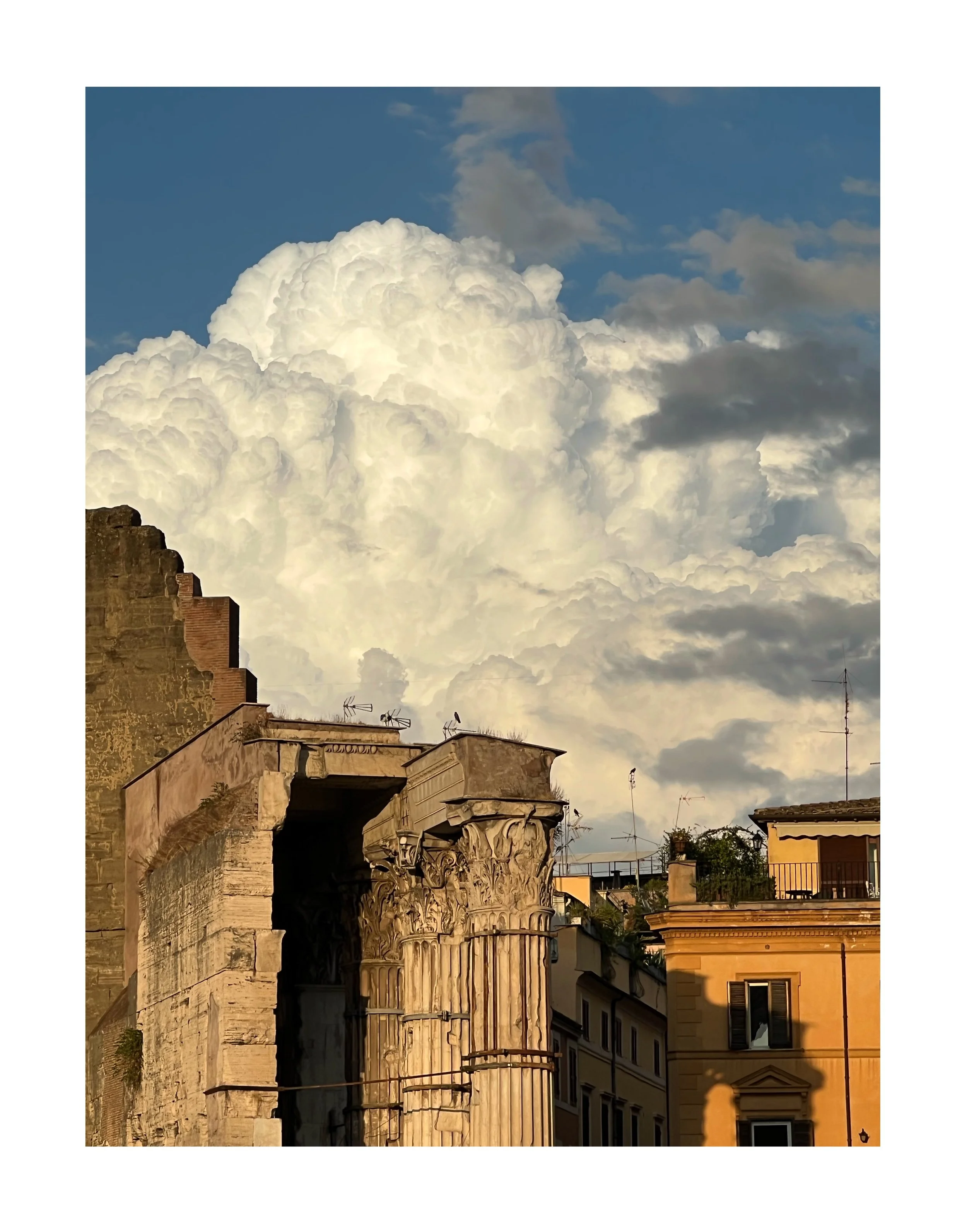 Ruines antiques avec colonnes ornées, bâtiments modernes sous un ciel avec de grands nuages blancs.