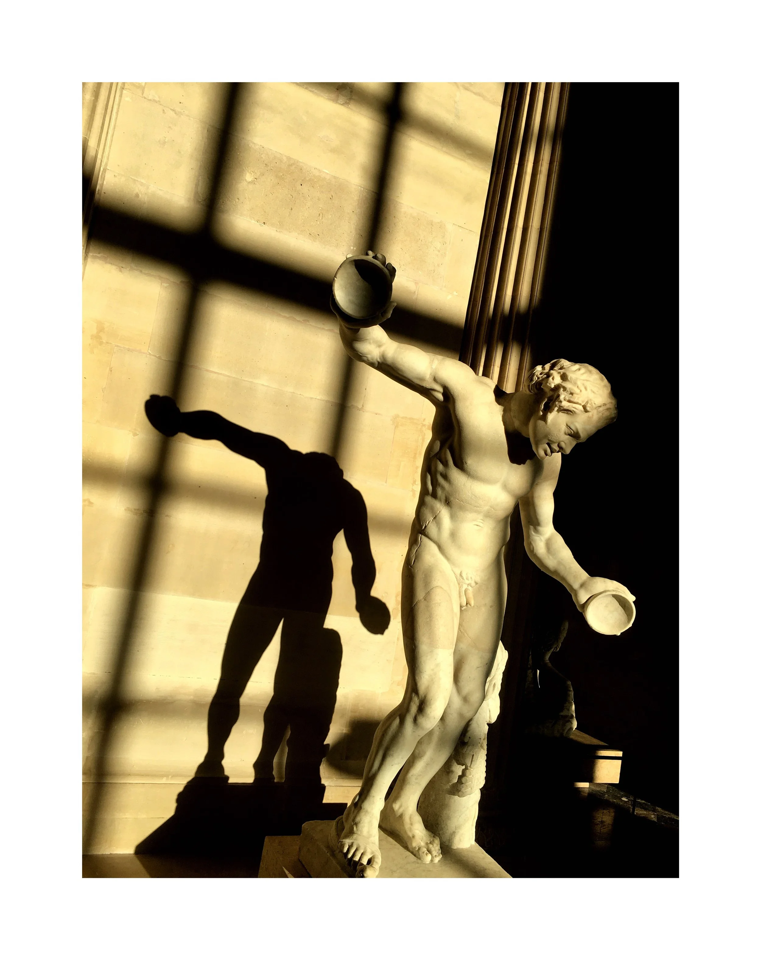 Statue de un homme nu avec un casque, tenant deux objets, avec une ombre de personne faisant du sport projetée sur le mur derrière lui.