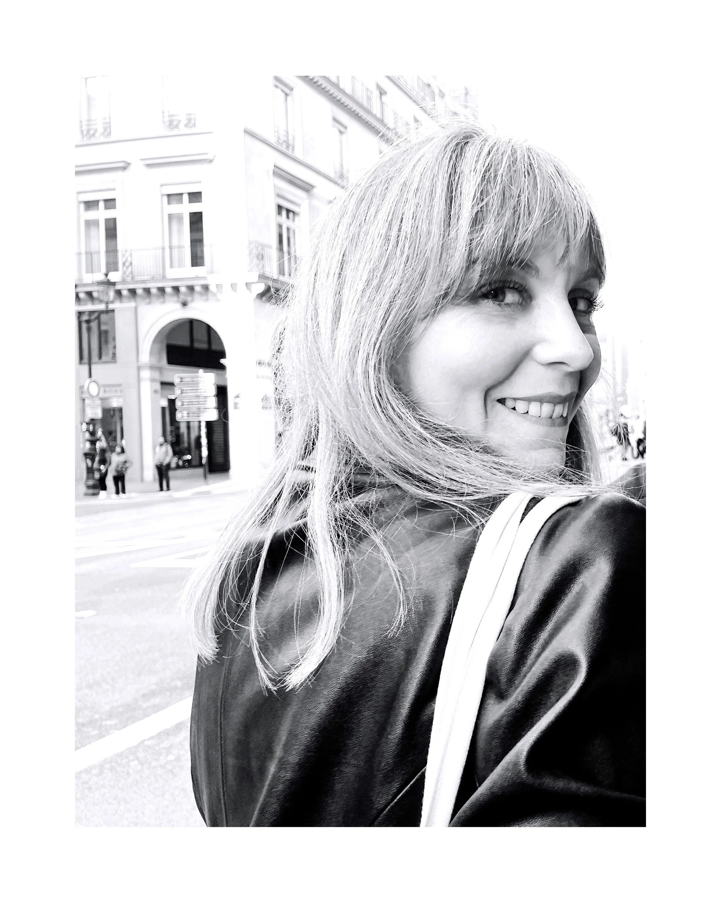 Une femme souriante aux cheveux longs et clairs, portant une veste en cuir, dans une rue urbaine. Fond de bâtiments historiques avec plusieurs personnes marchant et des panneaux de signalisation.
