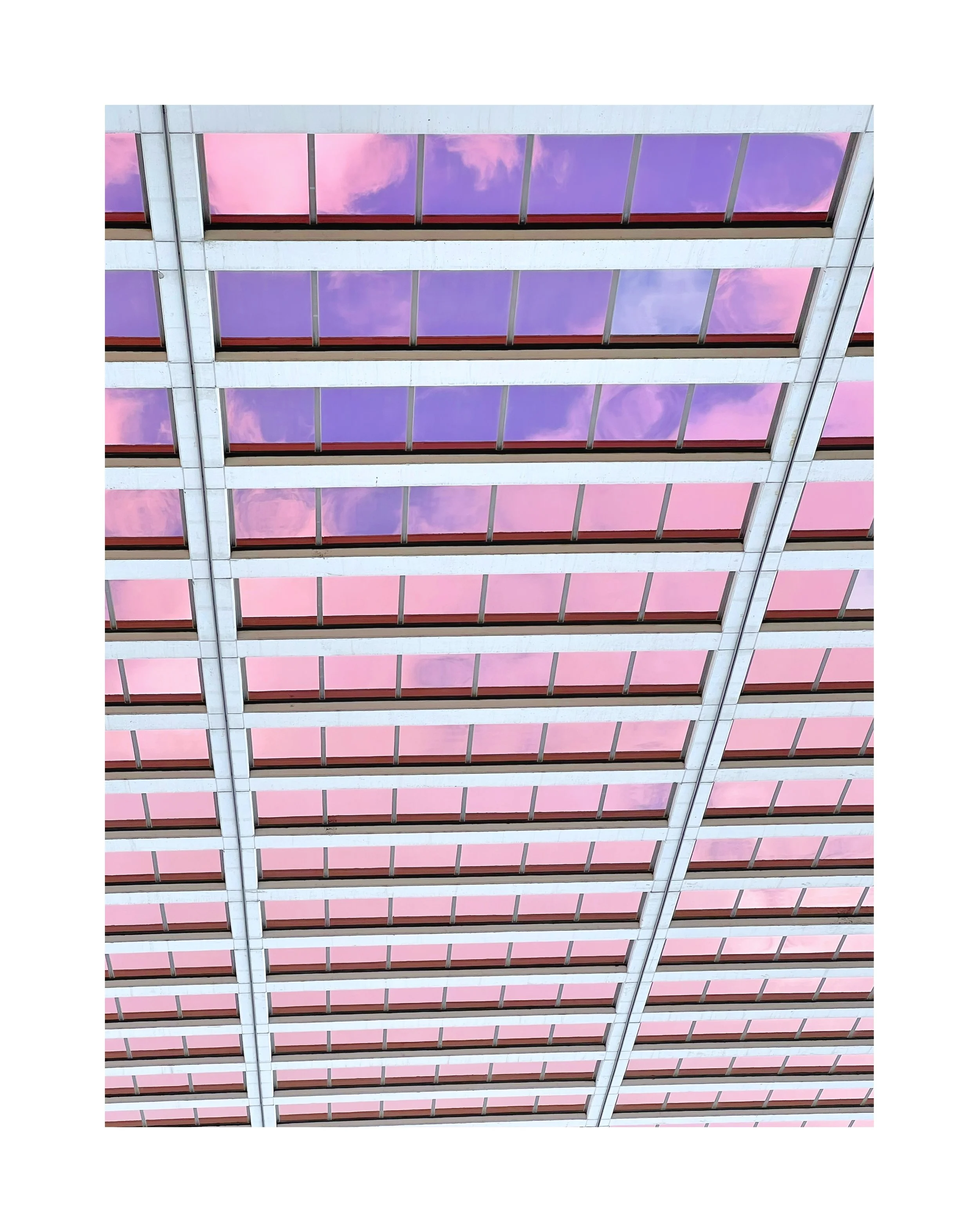 Vue du plafond d'un bâtiment avec des fenêtres en verre reflétant un ciel avec des nuages roses et violets.